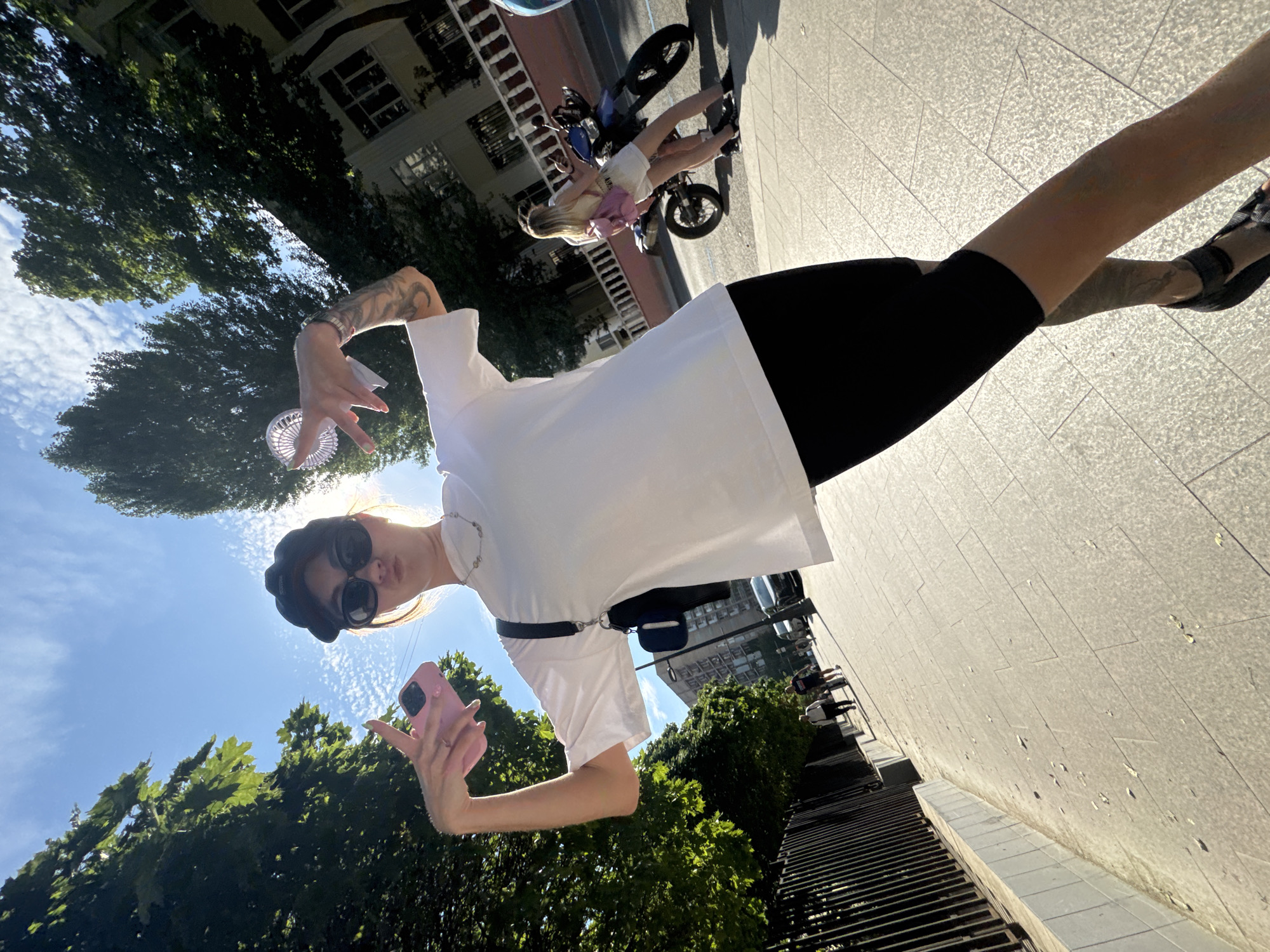 The image captures a young woman taking a selfie outdoors, likely in a city. She is wearing a white t-shirt, black shorts, sunglasses, and a black cap. She holds a pink smartphone in her right hand, and her left hand is extended, holding a small white portable fan. Her arm and torso are visible, with tattoos on her arm. She is wearing a necklace and a watch.
In the background, there are trees, a building, and a cobblestone sidewalk. Further in the distance, a motorcycle with two people on it is visible, suggesting a busy urban environment. The scene appears to be set during the daytime, with bright sunlight casting shadows, indicating it's likely sunny weather. The sky is blue with some clouds.
The overall mood of the image seems casual and relaxed, fitting for a sunny day outing. The location is identified as Moscow, Russia, but there are no explicit text elements or strong visual cues within the image to confirm this specifically. The focus is on the woman and her selfie-taking activity.