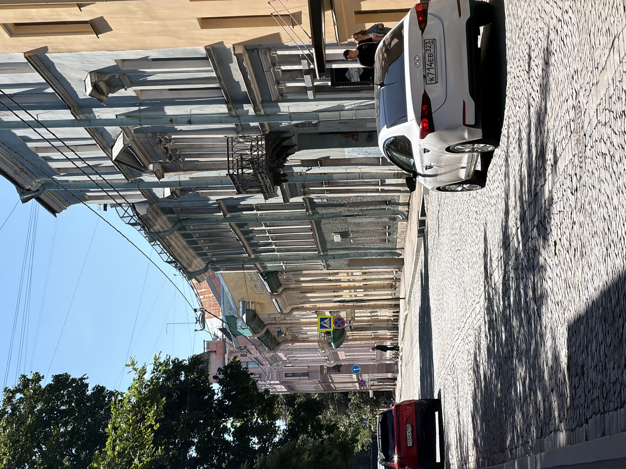 The image shows a sun-drenched, cobblestone street in Saint Petersburg, Russia. The street is lined with buildings exhibiting a mix of architectural styles and colors, ranging from pastel pink and yellow to muted blue and cream. The buildings feature classical detailing, with ornate window frames, cornices, and some balconies. A lush green tree is visible on the left side, casting dappled shadows across the road.
Two cars are present: a red car parked on the left and a white sedan driving away from the camera on the right. The white sedan has a Russian license plate that reads "K 714EB 323". In the background, several traffic signs are visible, including a pedestrian crossing sign and prohibition signs. A few people are walking on the sidewalk in the distance, one man standing near a building entrance. The sky is clear and blue. The overall impression is of a bright, warm day in an old European city.