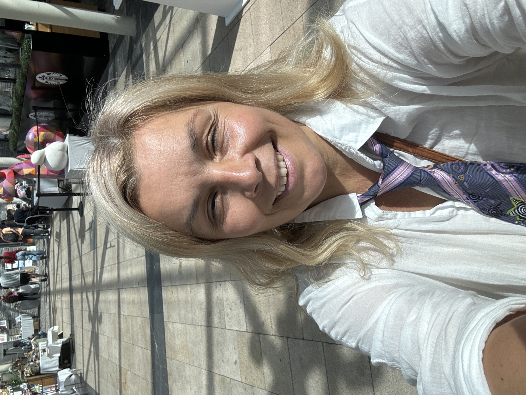 A blonde woman is taking a selfie in a well-lit outdoor pedestrian area. She is wearing a white button-down shirt and a patterned tie. Her eyes are closed, and she is smiling. In the background, people are walking and browsing at market stalls. The environment appears to be a modern shopping or market district with a tiled floor and architectural elements suggestive of a covered walkway. The image is taken during the day, with sunlight casting shadows on the ground. There is no information in the image to indicate the location is Kilkenny, Ireland.