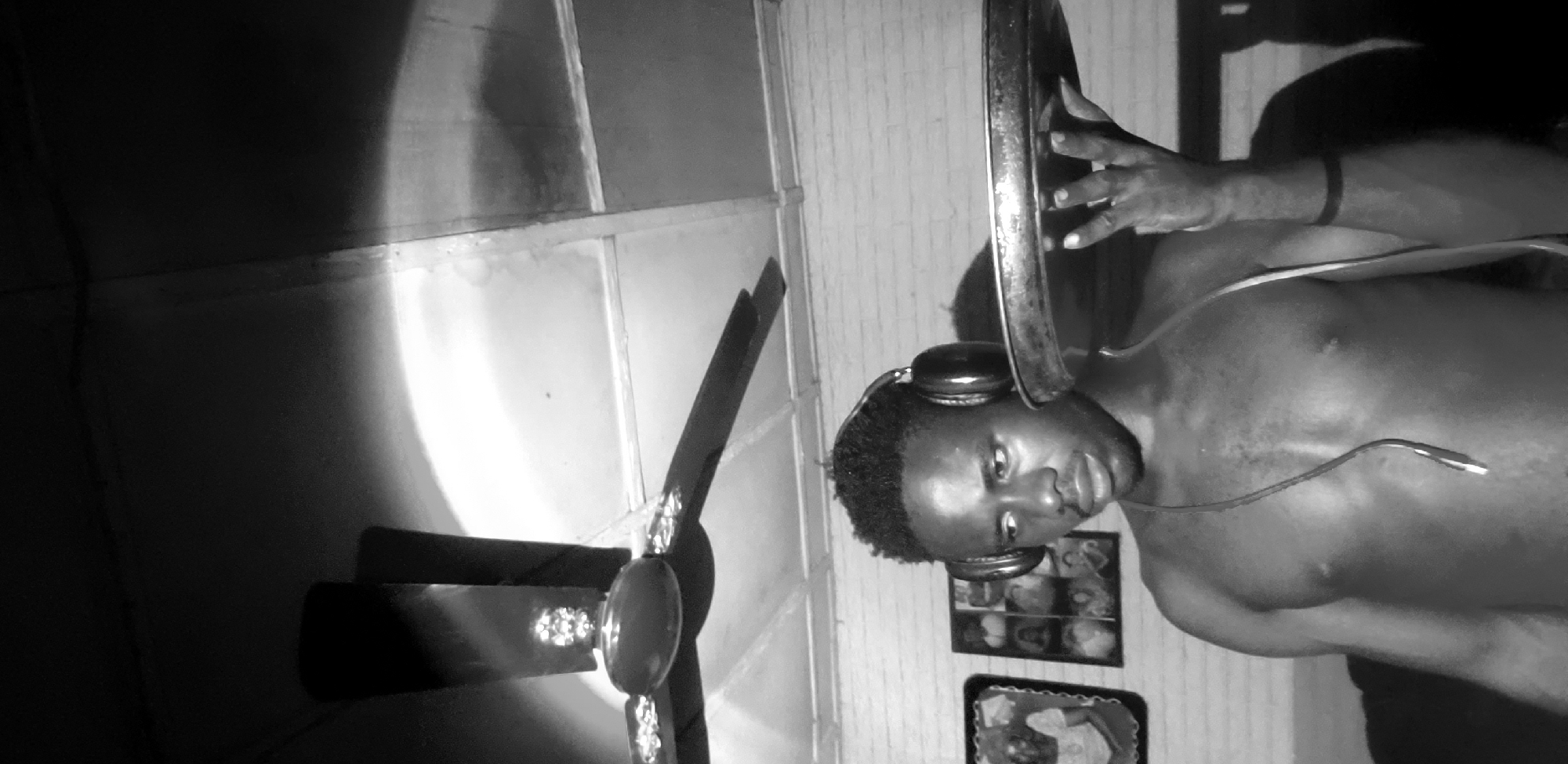 The image, captured in Narayi, Nigeria, is a medium shot in black and white, focusing on a bare-chested young Black male wearing headphones. He is holding a large, round, flat metallic object, possibly a tray or a large plate, with his right hand. The object is positioned in front of his chest and slightly to the right. His gaze is directed downwards and to his left. Behind him, a ceiling fan with three blades is visible, casting a shadow on the white, brick-like textured wall. To his left, on the wall, are two framed pictures. The lighting is dramatic, with a strong light source creating bright highlights and deep shadows, particularly on the ceiling and the subject's skin. The environment appears to be an indoor space, possibly a room.