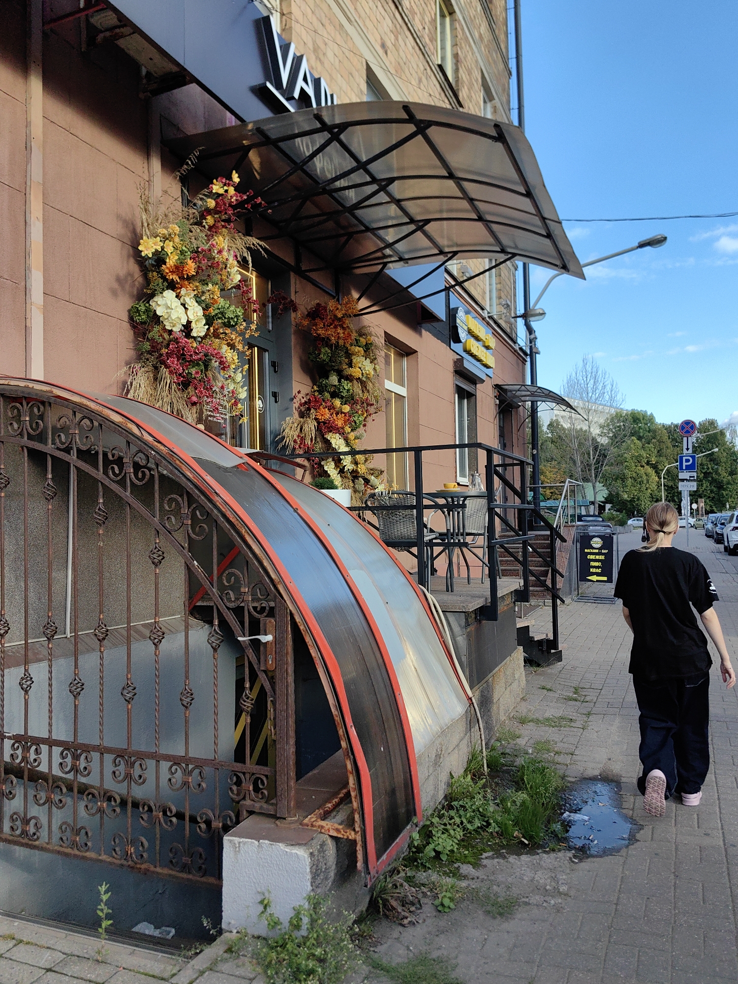 The image shows a street scene in Minsk, Belarus. In the foreground, a person with blonde hair pulled back in a ponytail, wearing a black t-shirt and black pants, walks away from the camera on a brick sidewalk. The person is wearing pink sneakers. To the left of the sidewalk is a metal fence with decorative scrollwork, enclosing a downward-sloping area with a covered entrance. The entrance has a translucent, curved roof with red trim. The building facade is brick and a light reddish-brown color. Above the entrance is a black awning, and a sign with the letters "VAN" in white is visible. Flower arrangements in autumnal colors are displayed on the building's facade. Further down the street, there is a street lamp, more trees, and parked cars. A blue and white sign with a parking symbol is also visible. The sky is clear and blue.