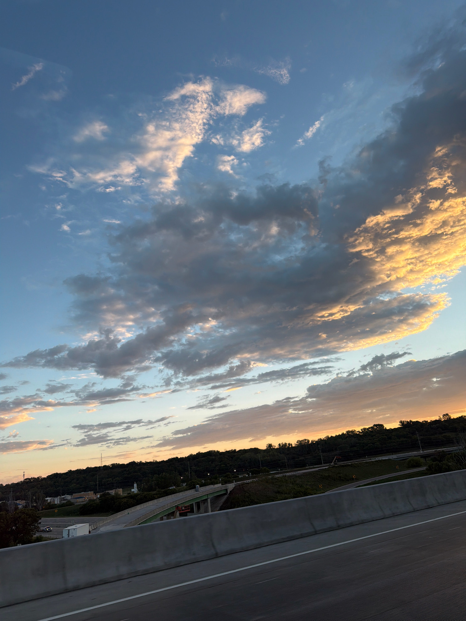 This is a photograph of a sunset over a highway and surrounding landscape. The sky is filled with dramatic clouds, some of which are illuminated by the setting sun, creating a warm, golden glow. The clouds range in color from white and pink to dark gray. Below the sky, a tree-covered hill forms the horizon. A highway with a concrete barrier runs through the foreground, with a bridge and some buildings visible in the distance. The overall impression is one of natural beauty and a sense of transition as day turns into night.