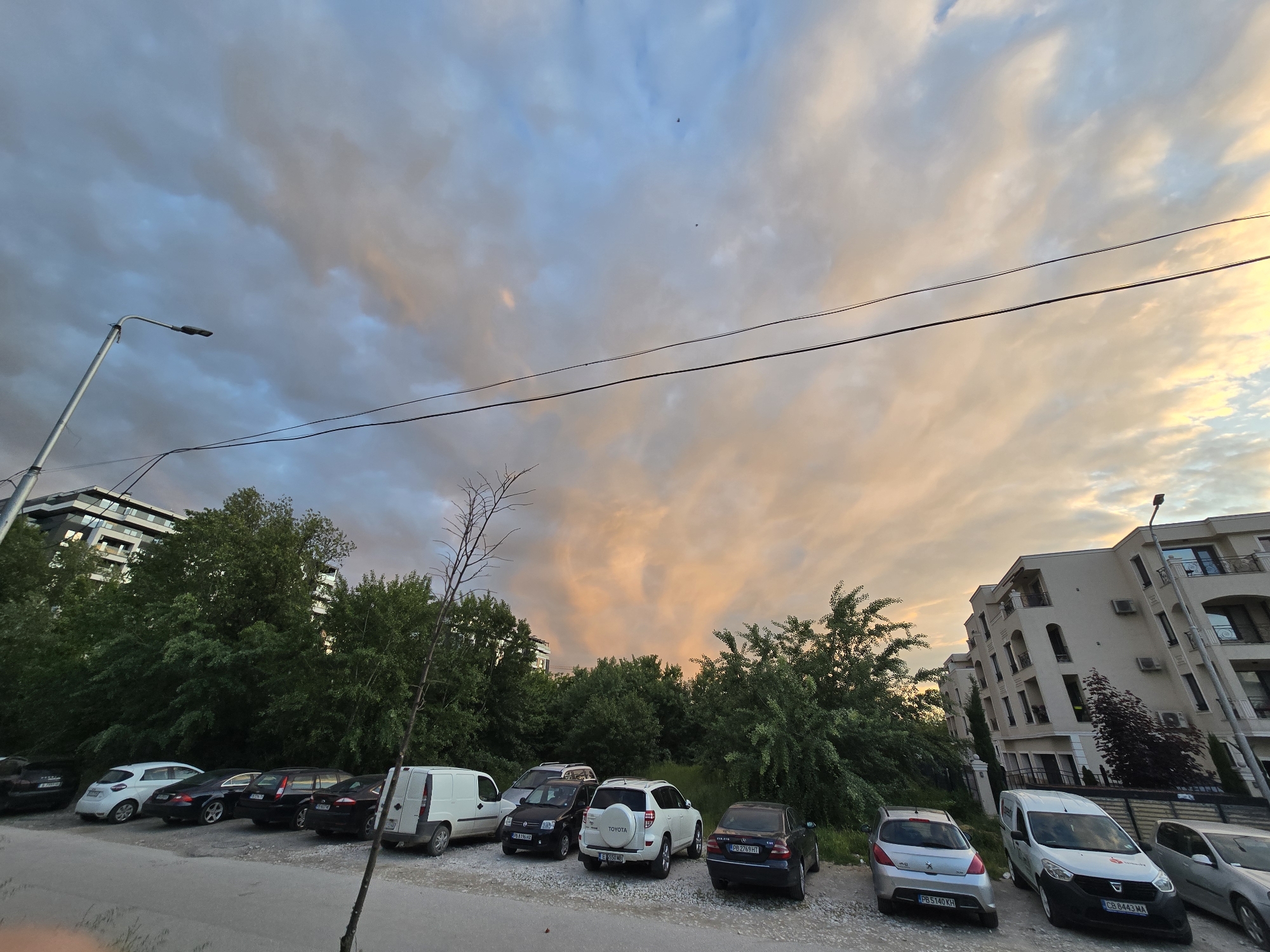 The image depicts a parking lot with a row of cars parked along a curb. Beyond the parking lot, there's a dense green area with trees and bushes. Buildings flank the scene on both sides, with a modern apartment building visible on the left and a multi-story residential building on the right. Above, the sky is filled with a mix of blue and orange-tinted clouds, suggesting either sunrise or sunset. Overhead wires run across the sky. A streetlight stands tall on the left side of the frame. The location appears to be an urban area, though the specific city and country are not discernible from the image.