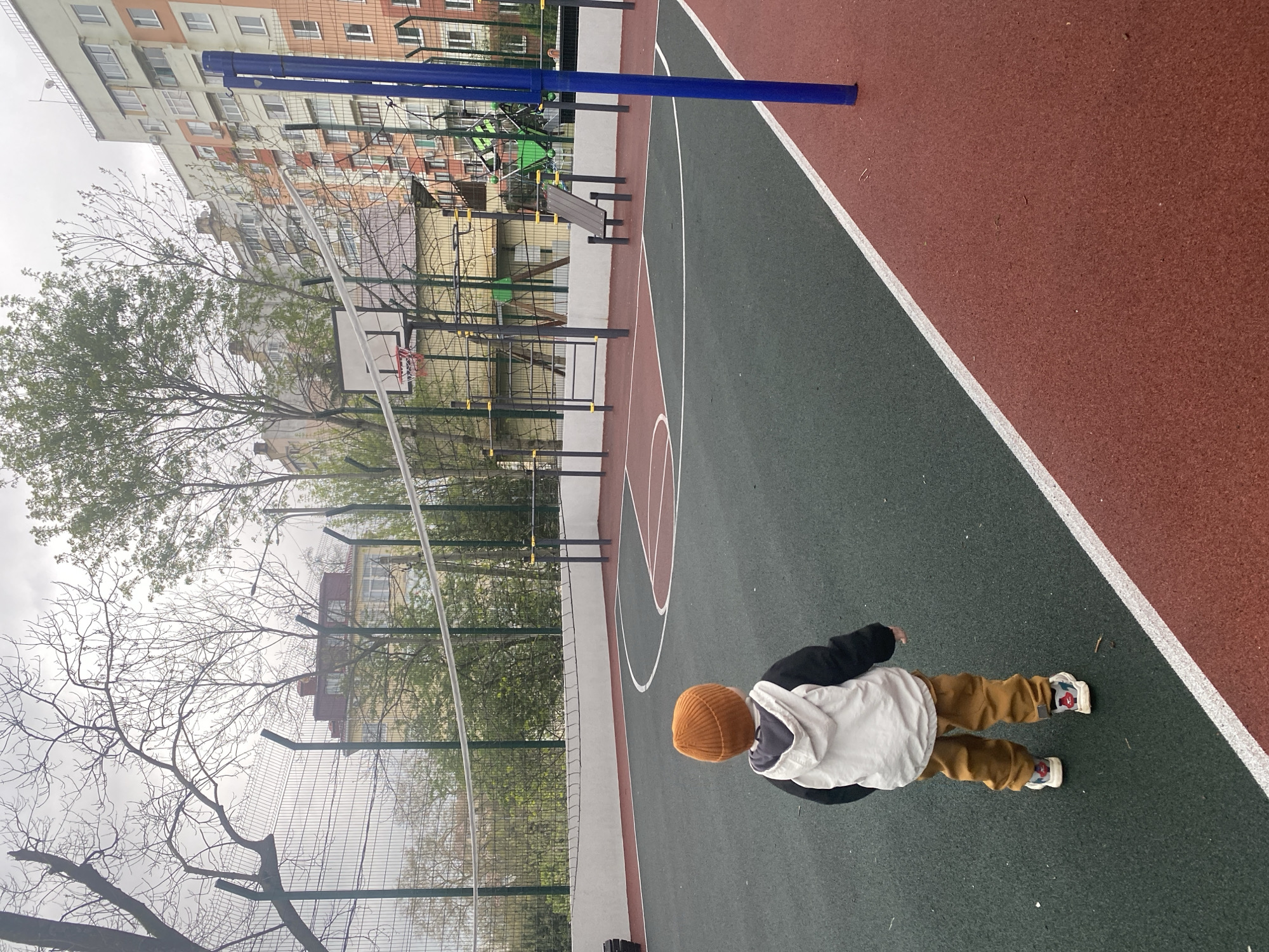 An overhead view captures a child walking on an outdoor sports court in what appears to be a residential area in Sochi, Russia. The court features a green playing surface delineated by white lines, including a free-throw line arc and a center circle arc, with an adjacent reddish-brown section.
The child, seen from behind, is dressed in a brown or orange knit beanie, a white and black hoodie or jacket, and brown or khaki trousers. They wear white sneakers with red and blue accents and appear to be walking towards a playground or outdoor gym area.
In the background, a basketball hoop with a white backboard and a red net is visible. Further back, various metal structures for exercise or play, some with green elements, are arranged within a fenced enclosure. Two tall blue poles stand prominently on the court, one closer to the child and another in the distance. Beyond the play area, multi-story residential buildings with orange/peach and white facades are visible under an overcast sky, suggesting a cloudy or muted daylight. Bare trees with some budding green leaves line the perimeter, indicating late winter or early spring. No visible text is present in the image.