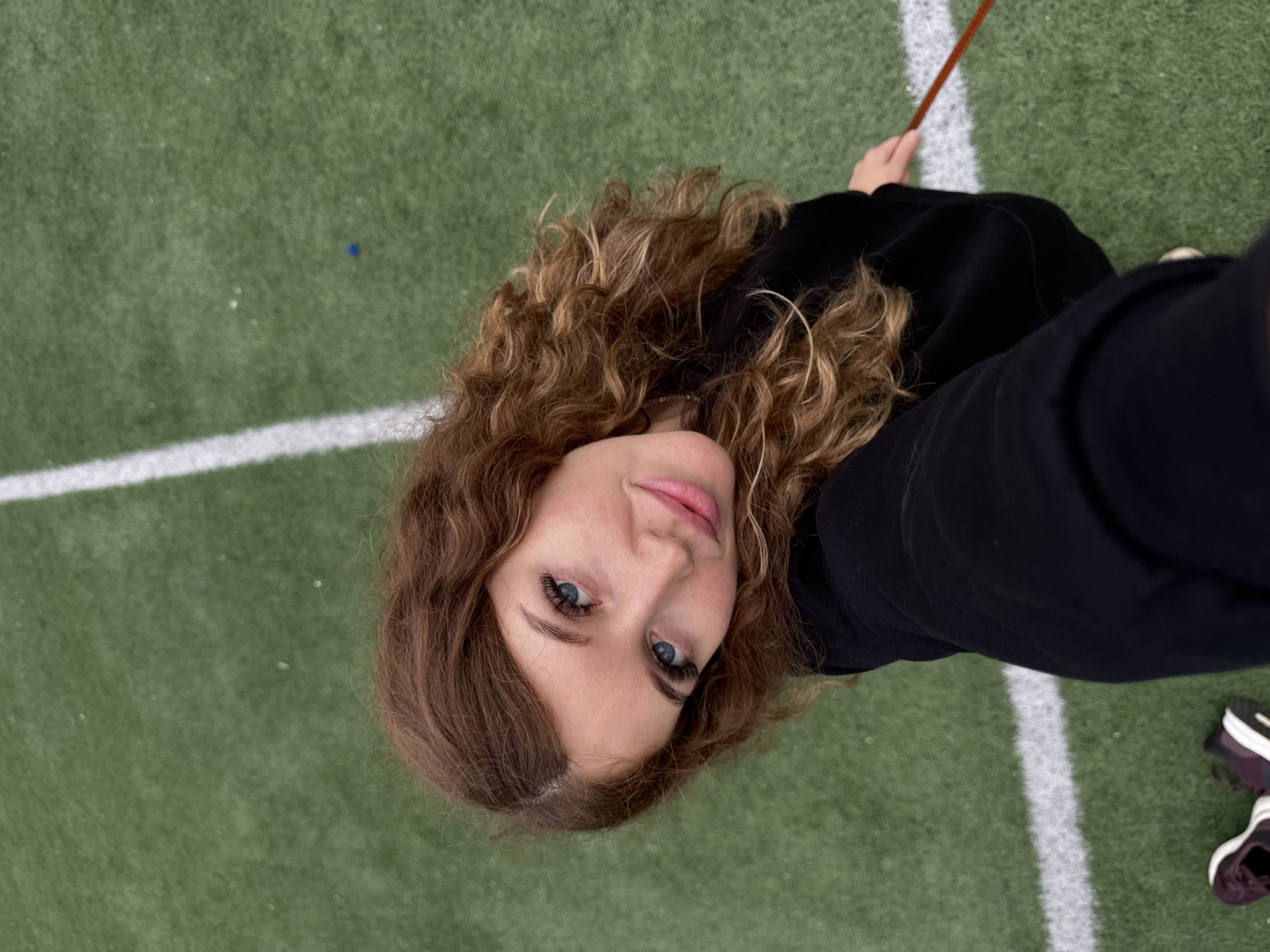 A young woman with long, curly auburn hair and blue eyes is taking a selfie from a high angle. She is wearing a black hoodie and has on what appears to be eyelash extensions. Her face is turned towards the camera, and she has a slight smile. She is standing on a green artificial turf field with white lines, characteristic of a sports field. The field is the likely location. The woman's shoes, dark purple sneakers, are visible at the bottom left of the frame. Her right hand is holding a thin, reddish-brown stick or wand. The overall lighting suggests an overcast day. The background is a blur of the green turf and white lines.