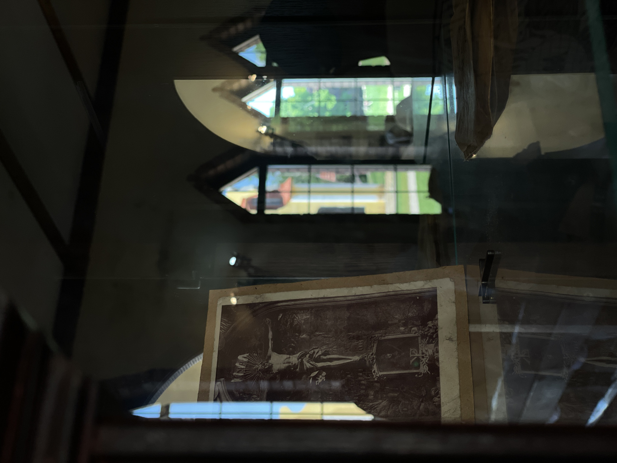 The image shows a sepia-toned photograph of the crucifixion of Jesus Christ, displayed inside a glass case. The photograph is old and appears to be mounted on cardboard. The background of the photograph depicts an ornate architectural setting. The glass case reflects the exterior, revealing arched windows with stained glass, through which a sunlit garden and building can be seen. The setting suggests an indoor display, possibly in a museum or historical site, located within the Літвенскі сельскі Савет, Belarus. The lighting is dim inside the display case, with highlights on the glass and the photograph. There are no people visible in the immediate foreground or interacting with the photograph. The overall scene is still and contemplative, focusing on the religious artifact.