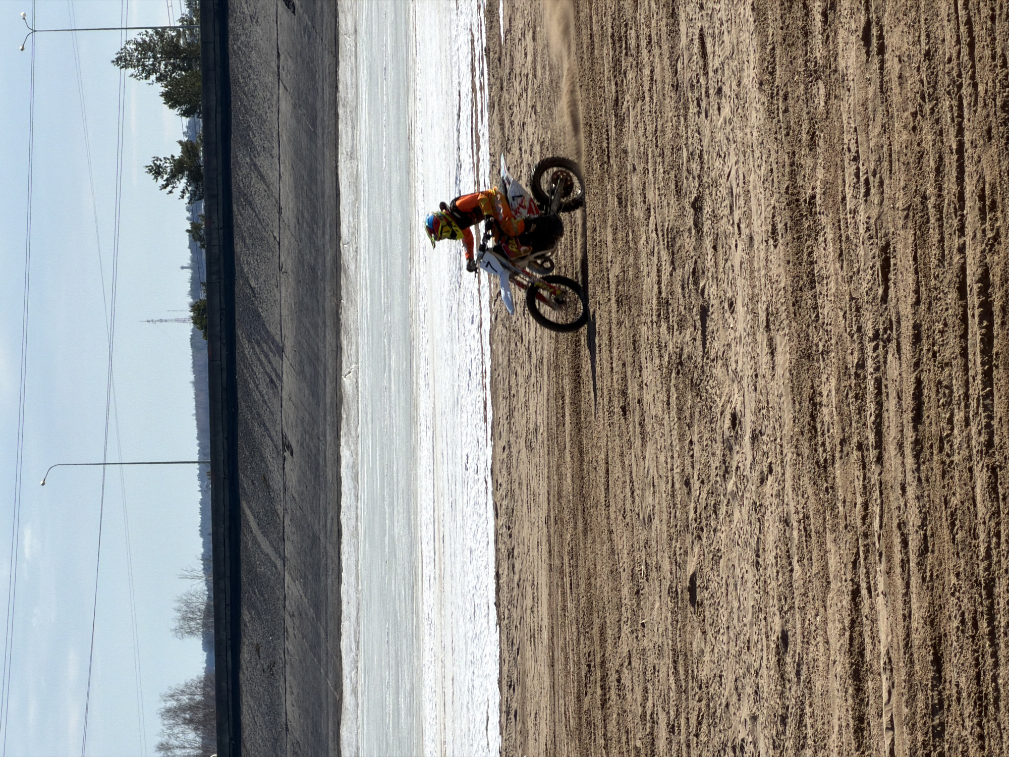 A person is riding a dirt bike on a dirt track next to a canal in the city of Chaikovsky, Russia. The person is wearing a helmet and riding gear. The canal is made of concrete and is filled with water. Trees and lampposts line the canal.