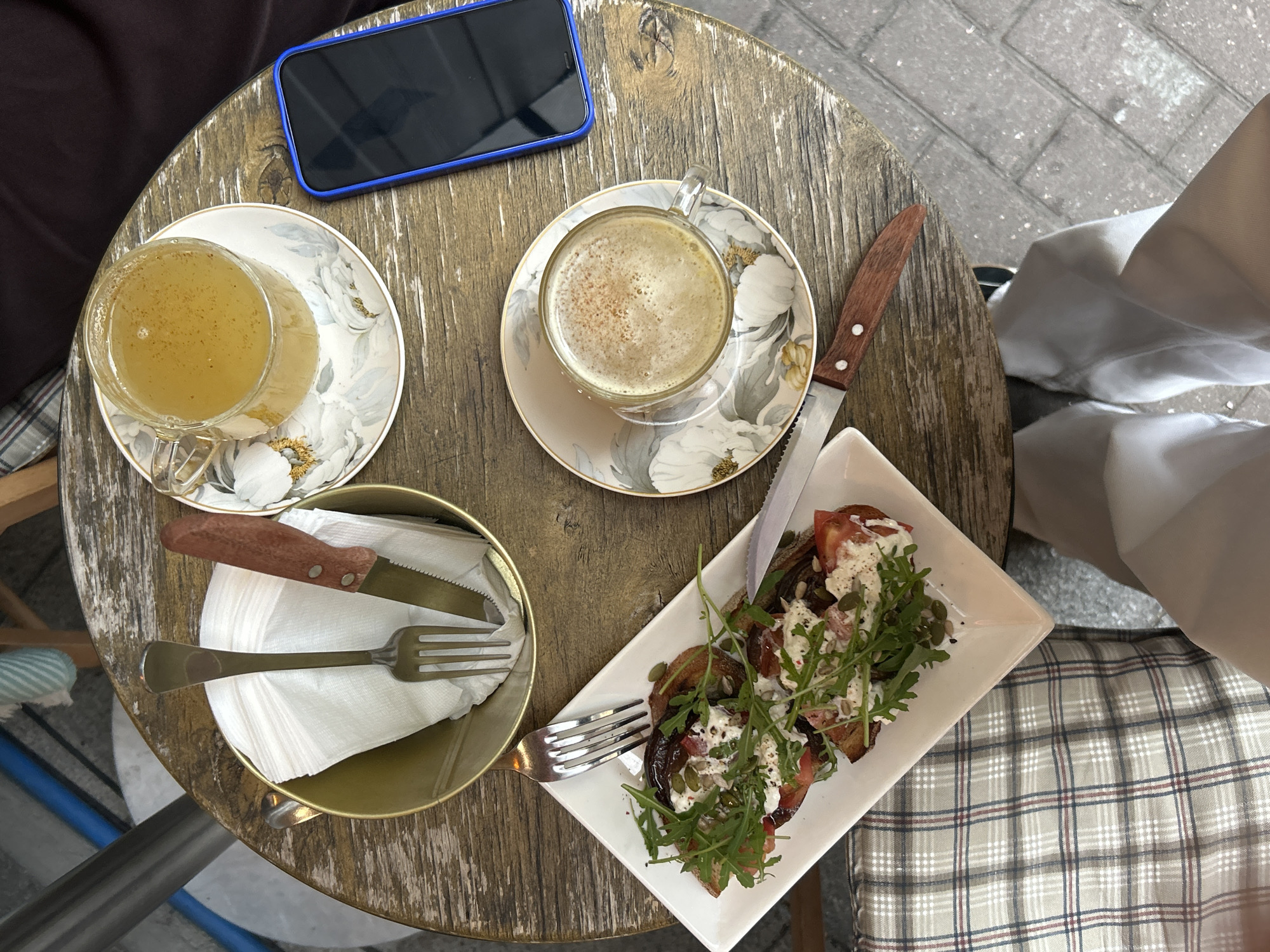 A top-down view captures a casual outdoor cafe setting, presenting a table with remnants of a meal and personal effects. A smartphone rests near the top left, adjacent to a teacup filled with a pale liquid, seated on a matching saucer. Another teacup, containing a frothy beverage with a sprinkle of powder, also sits on a saucer to the right. A wooden-handled knife lies beside it. A rectangular plate holds a partially consumed bruschetta topped with greens and tomatoes, accompanied by a fork and another wooden-handled knife. A circular container holds folded napkins along with cutlery. The table itself has a weathered, wooden appearance. The scene also includes portions of people's clothing and a patterned seat cushion. The location, identified as Saint Petersburg, Russia, is based on visual elements of the photograph.