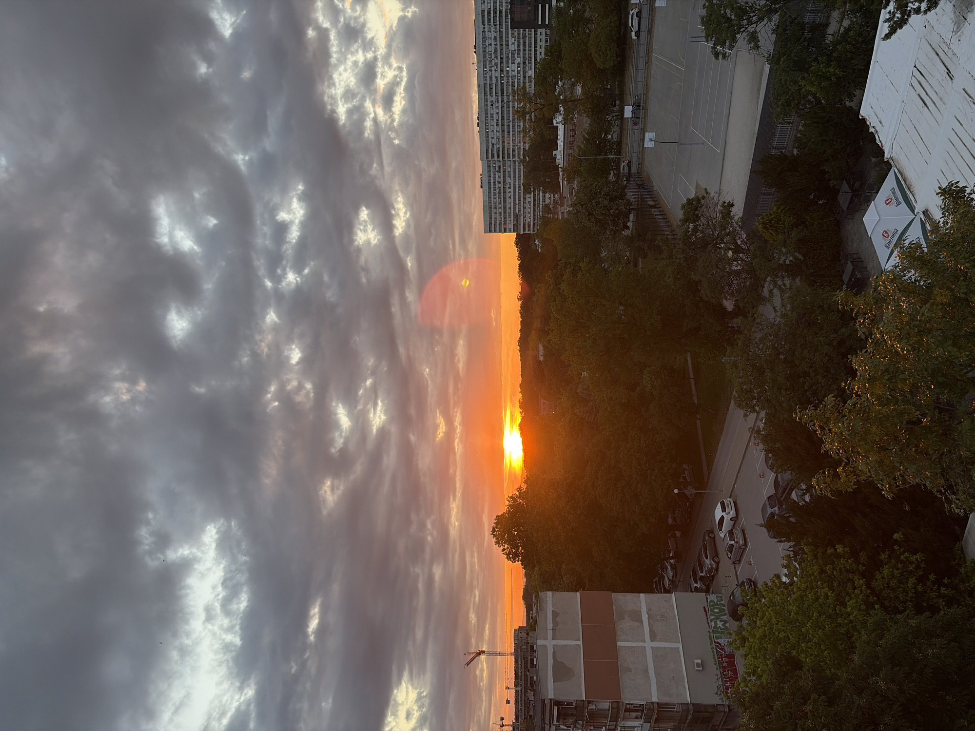 The image depicts a sunset over Plovdiv, Bulgaria. The sky is filled with clouds, illuminated by the setting sun. The sunset's orange glow contrasts with the grey clouds. Below the horizon, the image shows buildings, including residential complexes and what appears to be a construction site with a crane. Trees line the street. A basketball court and parked cars are visible.