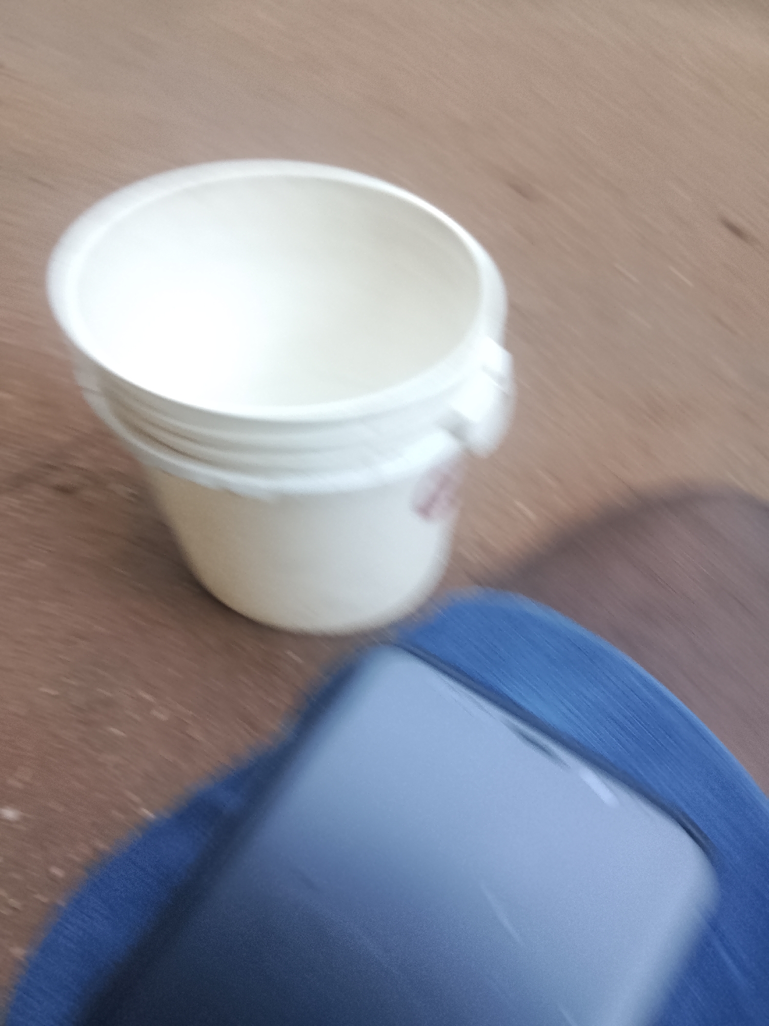 The media file shows a close-up, angled shot of a white plastic bucket with a rim. The bucket appears to be stacked, with at least two layers visible. It is situated on a wooden surface, possibly a table or floor. In the foreground, a blurred object, likely a smartphone, is placed on a blue fabric surface. The background is also blurred, with the wooden texture of the surface being the most discernible element. The lighting suggests an indoor setting, though no specific details about the environment are clear due to the blurriness. No people are visible in the frame. The overall impression is that of a casual, everyday scene, possibly a snapshot of items on a desk or workspace. The blurriness suggests movement during the capture of the image. There is no visible text in the image.
