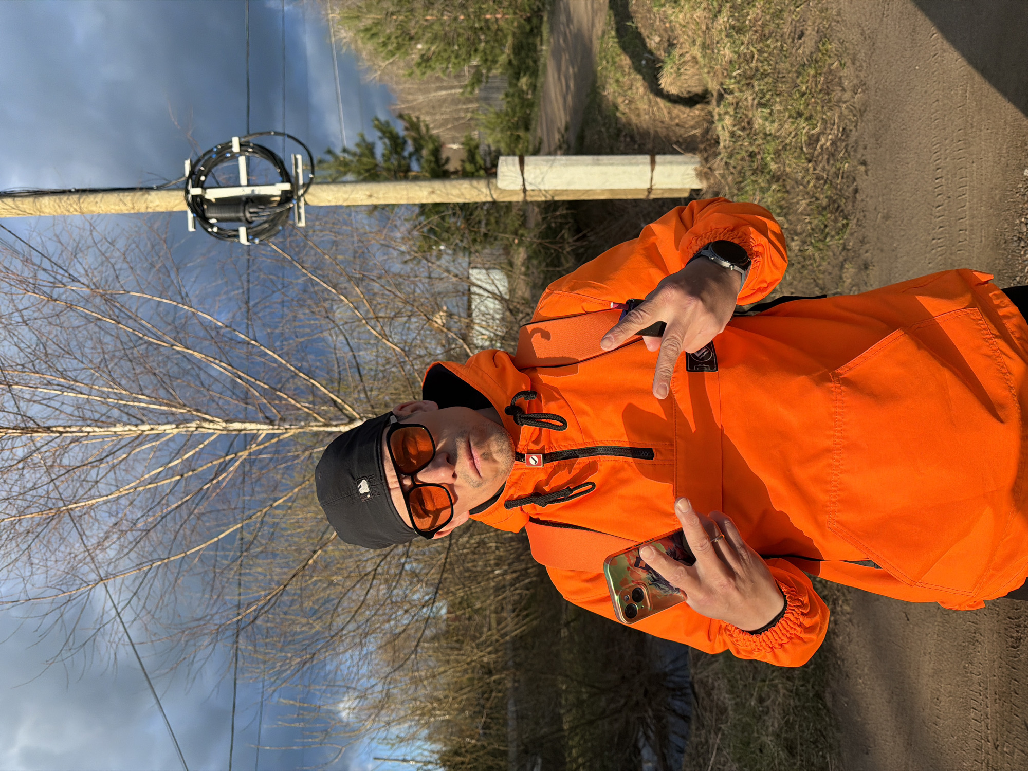 A man, appearing to be in his 20s-30s, is captured in an outdoor, vertical portrait. He wears a bright orange anorak jacket with black accents, a black beanie with a white "MLB" logo on the front, and orange-tinted sunglasses. He has light stubble and a neutral expression.
In his left hand, he holds a smartphone with a patterned case. His right hand is raised, forming a "peace" or "V" sign. A smartwatch with a metallic bezel is visible on his right wrist.
The setting appears to be a rural or suburban area in Chaikovsky, Russia. Behind the man, a wooden utility pole is visible, wrapped with bundled black cables, possibly fiber optic, held by white fasteners. Bare tree branches with dormant buds fill the mid-ground, suggesting late winter or early spring. In the distance, green conifer trees are visible, and a dirt path or road runs along the right side of the frame. The sky is partially cloudy, with patches of bright and darker, overcast skies. The lighting suggests it is daytime, possibly late afternoon, with some shadows evident.