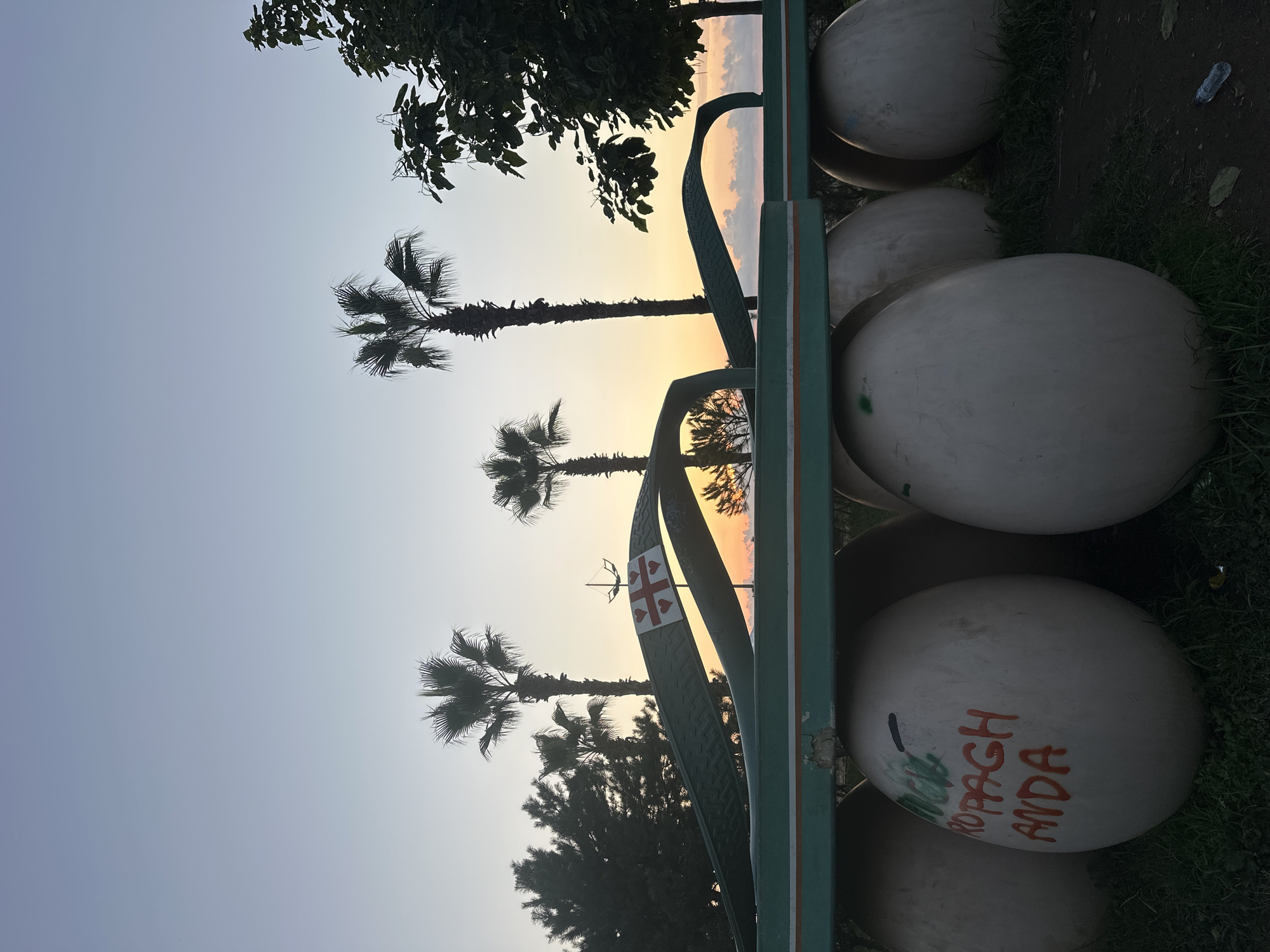 The image depicts a large, green, stylized flip-flop sculpture situated outdoors at dusk. The sculpture is elevated on several large, white, egg-shaped bases, some of which are marked with graffiti. In the background, several palm trees and other foliage are visible against a sky transitioning from a light blue at the top to orange and yellow hues near the horizon, indicating sunset. A portion of a flag with a red cross on a white background and red hearts within the cross segments is visible on the strap of the flip-flop sculpture. This sculpture is located at the "Alphabet Tower" in Batumi, Georgia, which is known for its unique architectural landmarks. The overall scene is calm and serene, characteristic of a coastal city at the end of the day.