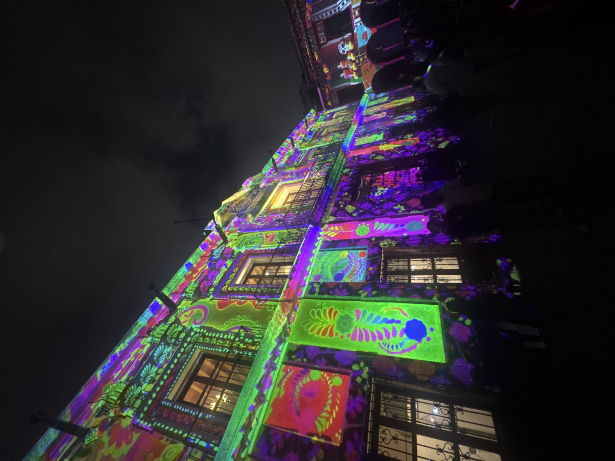 A low-angle shot depicts a multi-story building facade adorned with colorful light projections, likely taken at night in Puebla, Mexico. The building's architectural details are obscured by the vibrant light show, which uses various colors like green, purple, pink, and blue. The projections include floral motifs, patterns, and possibly traditional Mexican designs like skulls near the top.
The image seems to be taken from a pedestrian area, with glimpses of people visible at the top right of the frame. The sky is dark and overcast in the top left. The window designs on the building are ornate and suggest a historical building of some significance. The overall impression is of a festive or celebratory event.