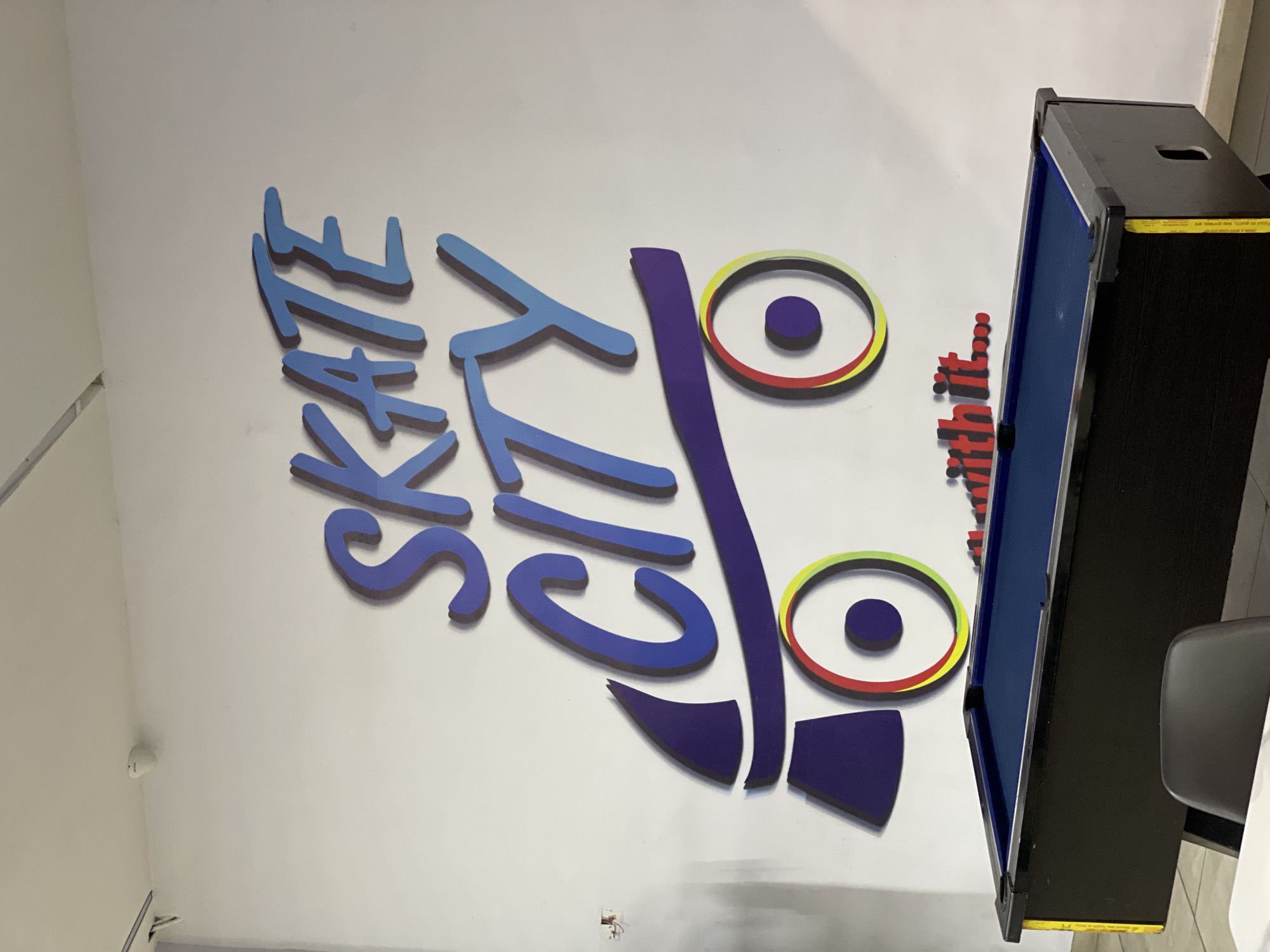 A photograph taken from a low angle shows a white wall with a large, colorful mural depicting the words "SKATE CITY" in large, stylized blue lettering. Below the text is a graphic of a roller skate with purple and blue accents and colorful wheels. To the right of the mural is a pool table with a blue felt top and black sides, a portion of a black chair is visible in the foreground. The word "with it..." is also visible in red lettering below the skate graphic. The setting appears to be an indoor recreational facility, possibly in Moba, Nigeria, given the prompt.