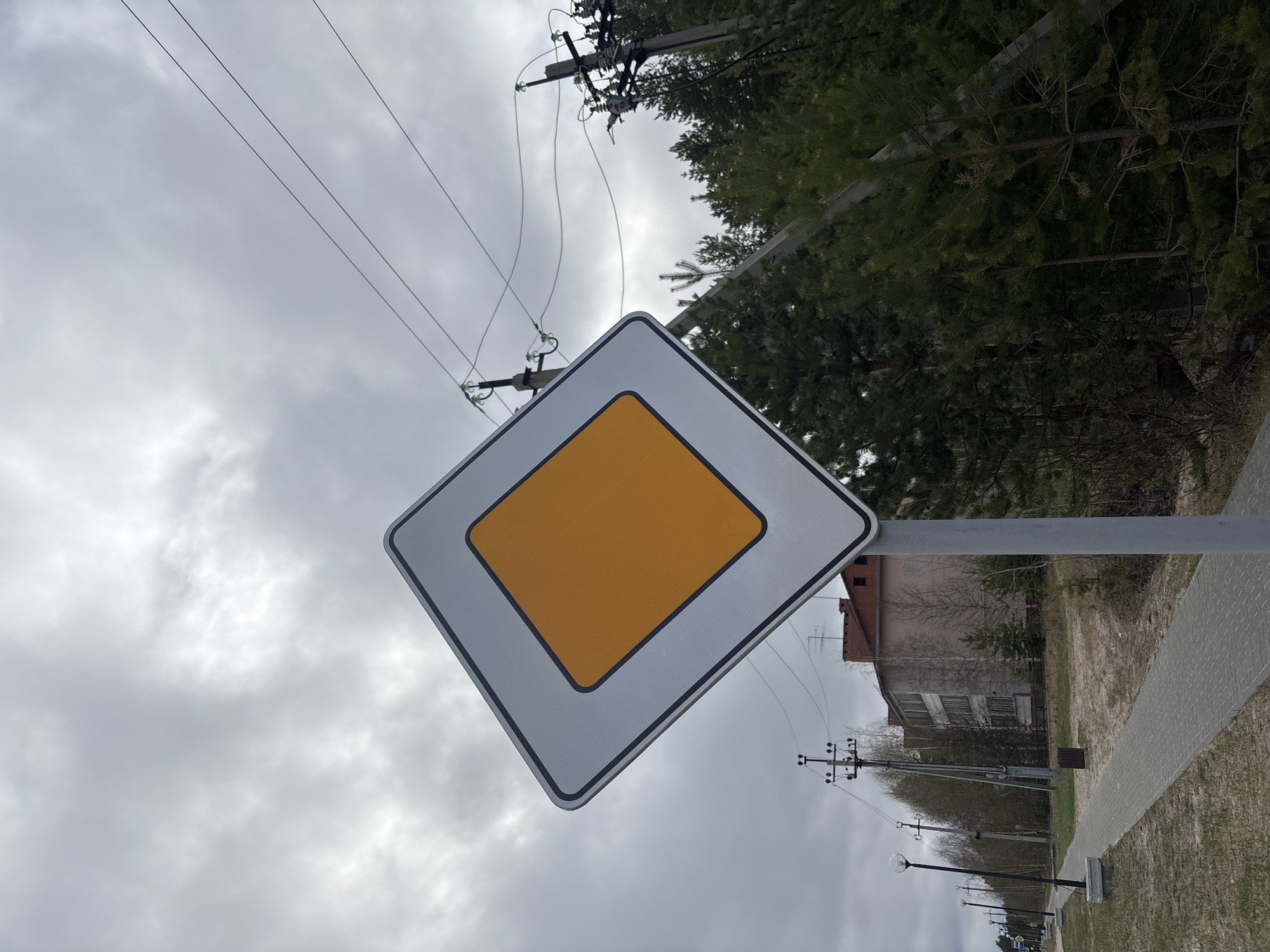 The image shows a diamond-shaped traffic sign with a yellow center and white border, indicating the main road or right-of-way. The sign is mounted on a metal pole against a cloudy sky. Electrical wires are visible in the upper portion of the frame, running between utility poles. In the background, there are trees, a building with a red roof, and a paved sidewalk. The scene is located in Верхнеказымский, Russia.