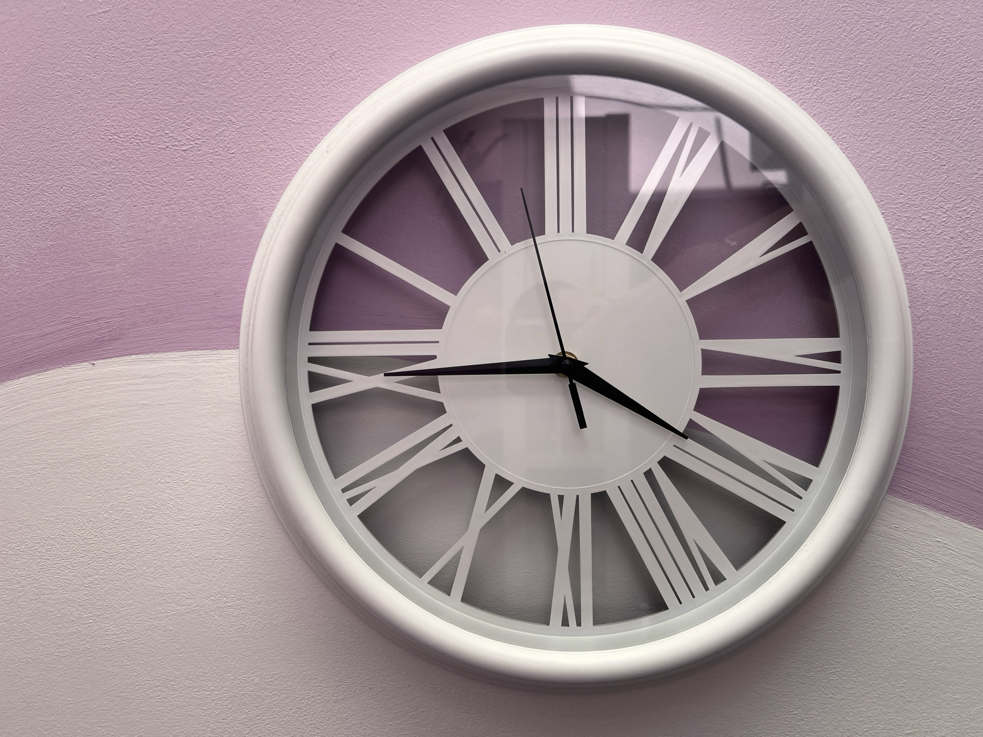 A white clock with Roman numerals is mounted on a light purple wall with a white stripe. The clock face is white and has black hands that point to approximately 4:27. The setting appears to be indoors. No people are visible. The location in Plovdiv, Bulgaria, is not evident from the image.