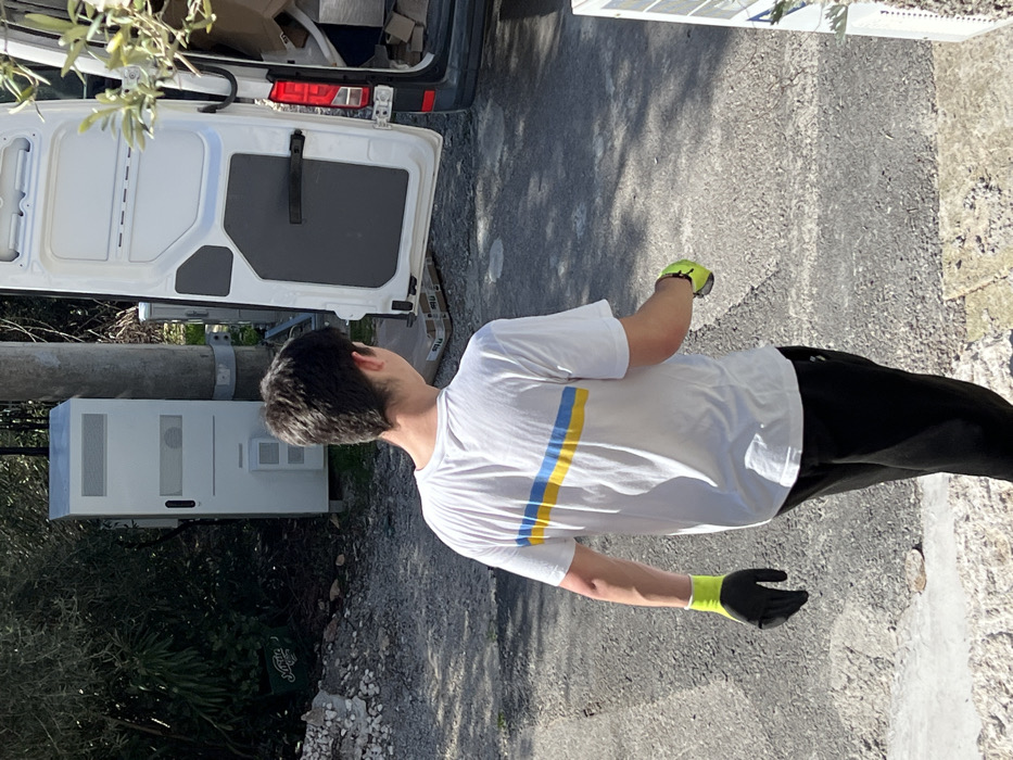 The scene is an outdoor setting in Bigova, Montenegro, illuminated by bright sunlight. A young male, viewed from behind, stands on an uneven surface of dirt, gravel, and asphalt, with piles of light-colored rubble visible on the left. He is wearing a white short-sleeved t-shirt with a horizontal blue stripe above a yellow stripe across the upper back, black athletic pants, and a black-and-neon-yellow work glove on his left hand. A matching glove is held in his right hand. To the right of the individual, a white panel van is parked with its rear doors fully open, revealing a dark grey interior containing cardboard boxes and other packed items. On the ground directly behind the van, several flat, rectangular packages with an "M" logo are visible. To the left of the van, a grey utility pole stands next to a tall, rectangular, light-grey utility cabinet equipped with ventilation grilles and access panels. Dense green foliage is present in the background, with a partially obscured green plastic crate visible on the far left.