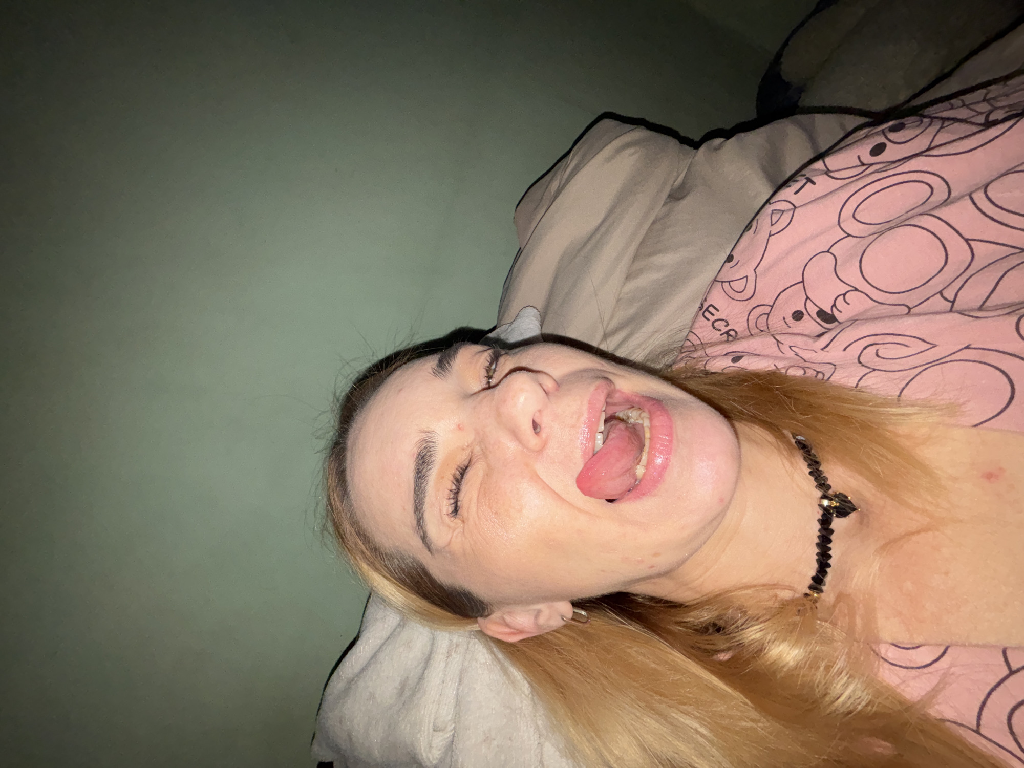 A woman is lying in bed, with her eyes closed and her tongue sticking out. She is wearing a pink shirt with a teddy bear pattern and a black beaded necklace. The room is dimly lit, and the walls are a muted green color. There are pillows and blankets visible in the background. The image appears to have been taken with a phone camera, possibly as a selfie. The woman's facial expression suggests she is playful or perhaps joking.