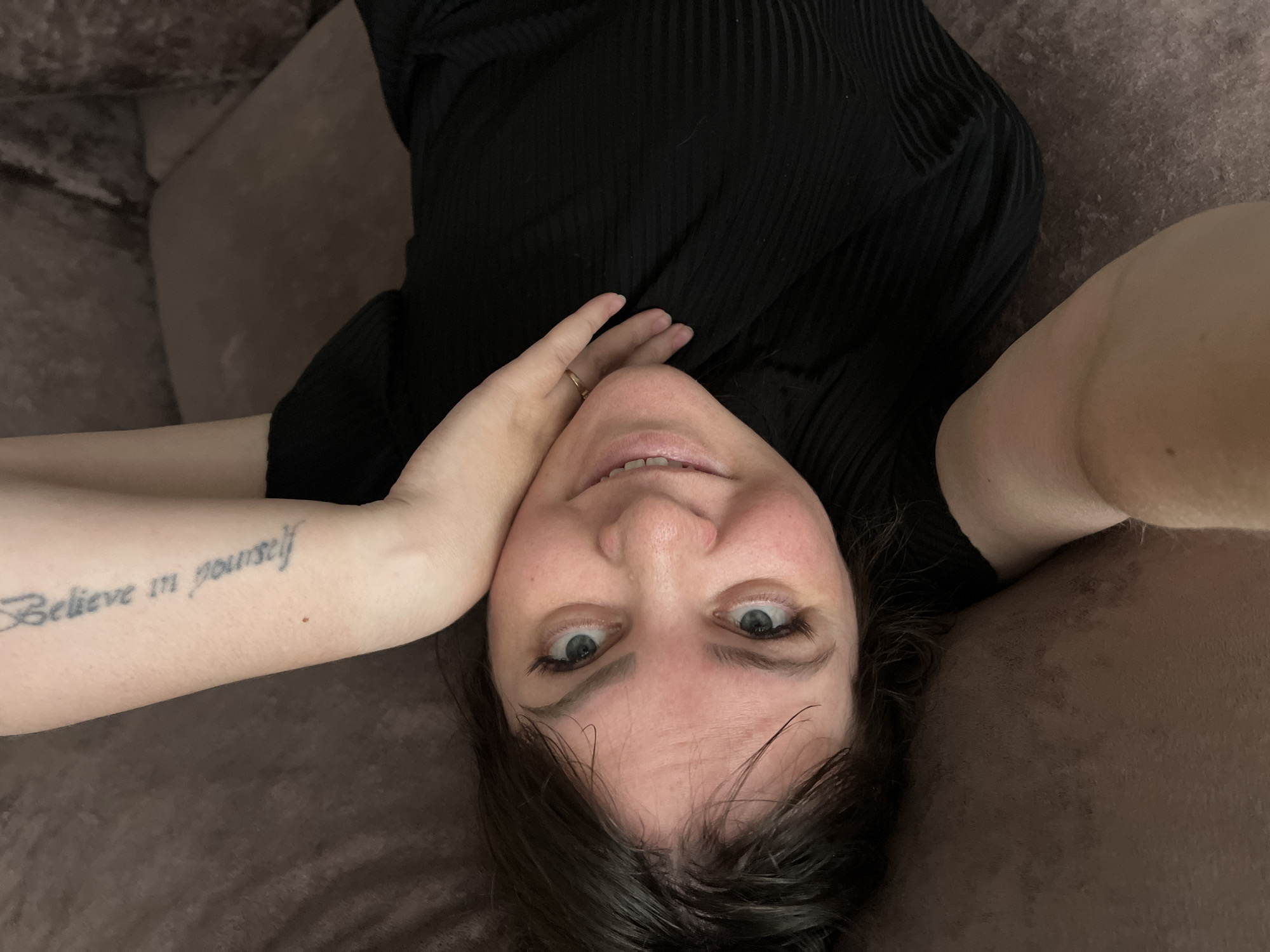 A woman is lying on a brown couch. She is wearing a black shirt. She has a tattoo on her left forearm that says "Believe in yourself." She is looking at the camera with her arms to either side of her face. The location is not discernible from the image.