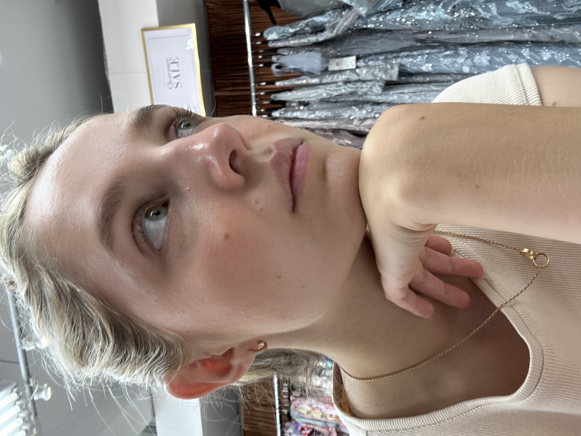 A close-up, eye-level, medium shot captures a young Caucasian woman with blonde hair pulled back, resting her chin on her hand as she looks to her right, out of frame. Her head is tilted slightly, and her eyes are open. The background features racks of sparkly dresses, suggesting a retail environment, with a "Clearance SALE" sign visible above. The lighting is bright, and the overall scene appears to be indoors, possibly in a clothing store in New York City.