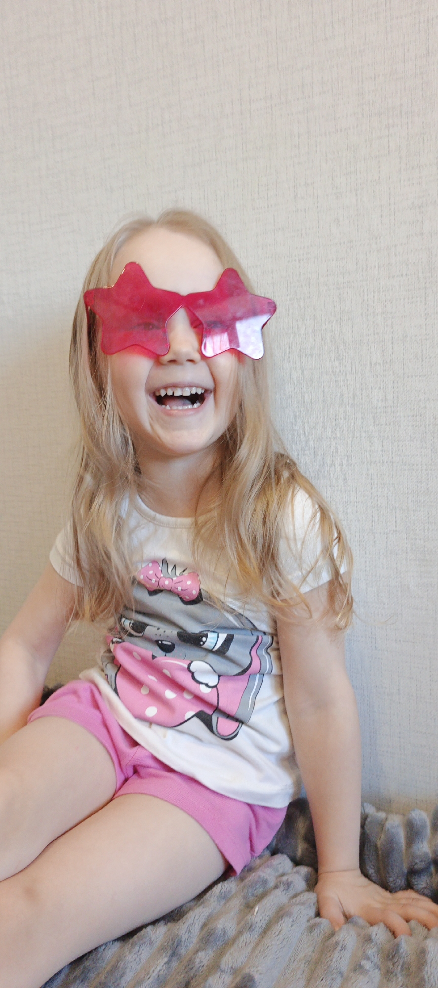 A young girl with blonde hair is laughing while wearing pink star-shaped sunglasses. She is wearing a white t-shirt with a cartoon cat design and pink shorts. She is sitting on a gray, textured blanket or cushion. The background is a plain, light-colored wall. The image may have been taken indoors. There is no indication of the location being Chaikovsky, Russia.