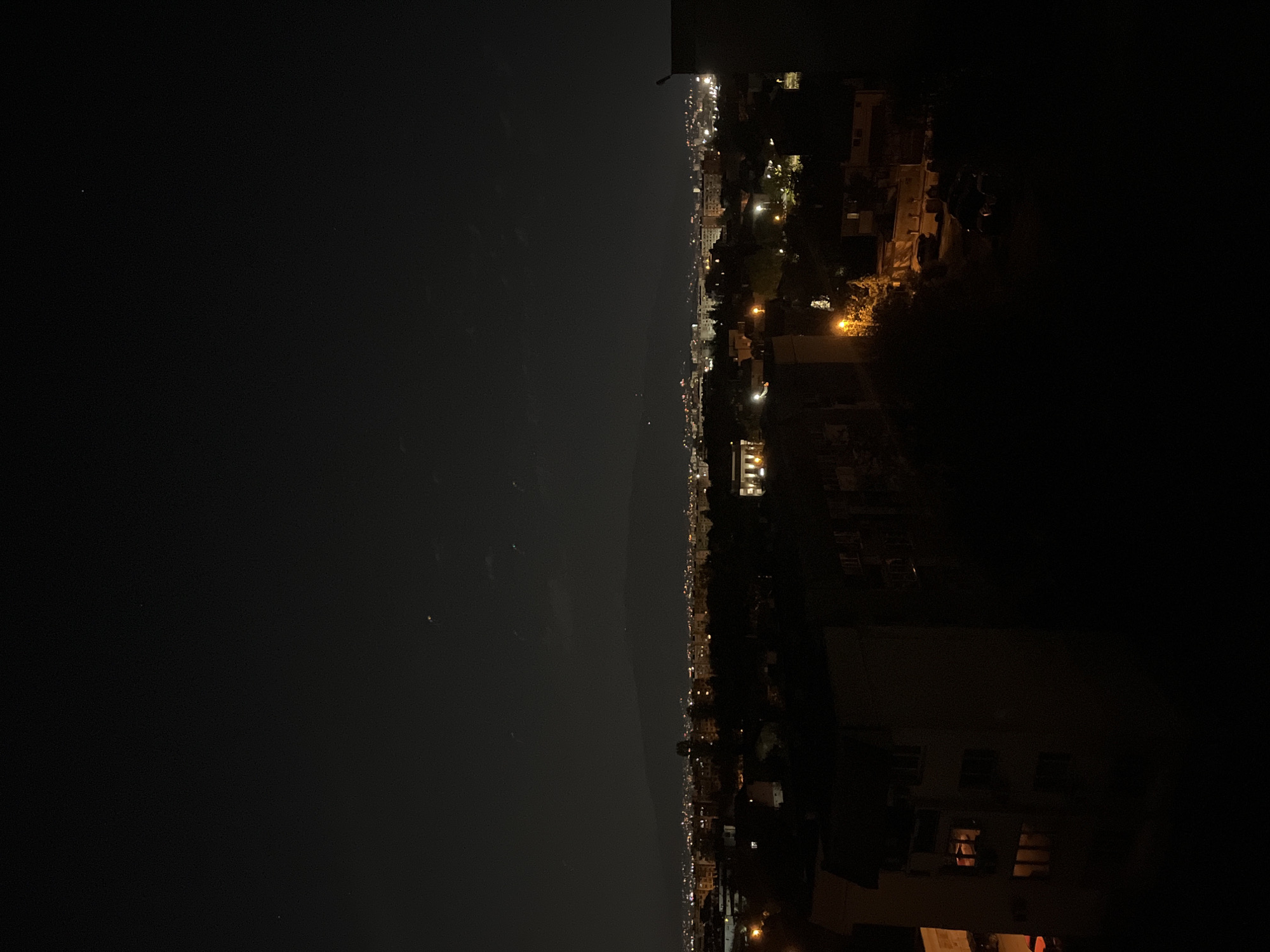 The image depicts a night view of Sofia, Bulgaria, as seen from a high vantage point. The left side of the image shows a dark sky with faint stars, while the right side reveals the cityscape illuminated by streetlights and building lights. The city lights form a linear pattern along the horizon, indicating roads and building structures. Buildings and other structures are also visible closer to the camera on the right-hand side of the image.