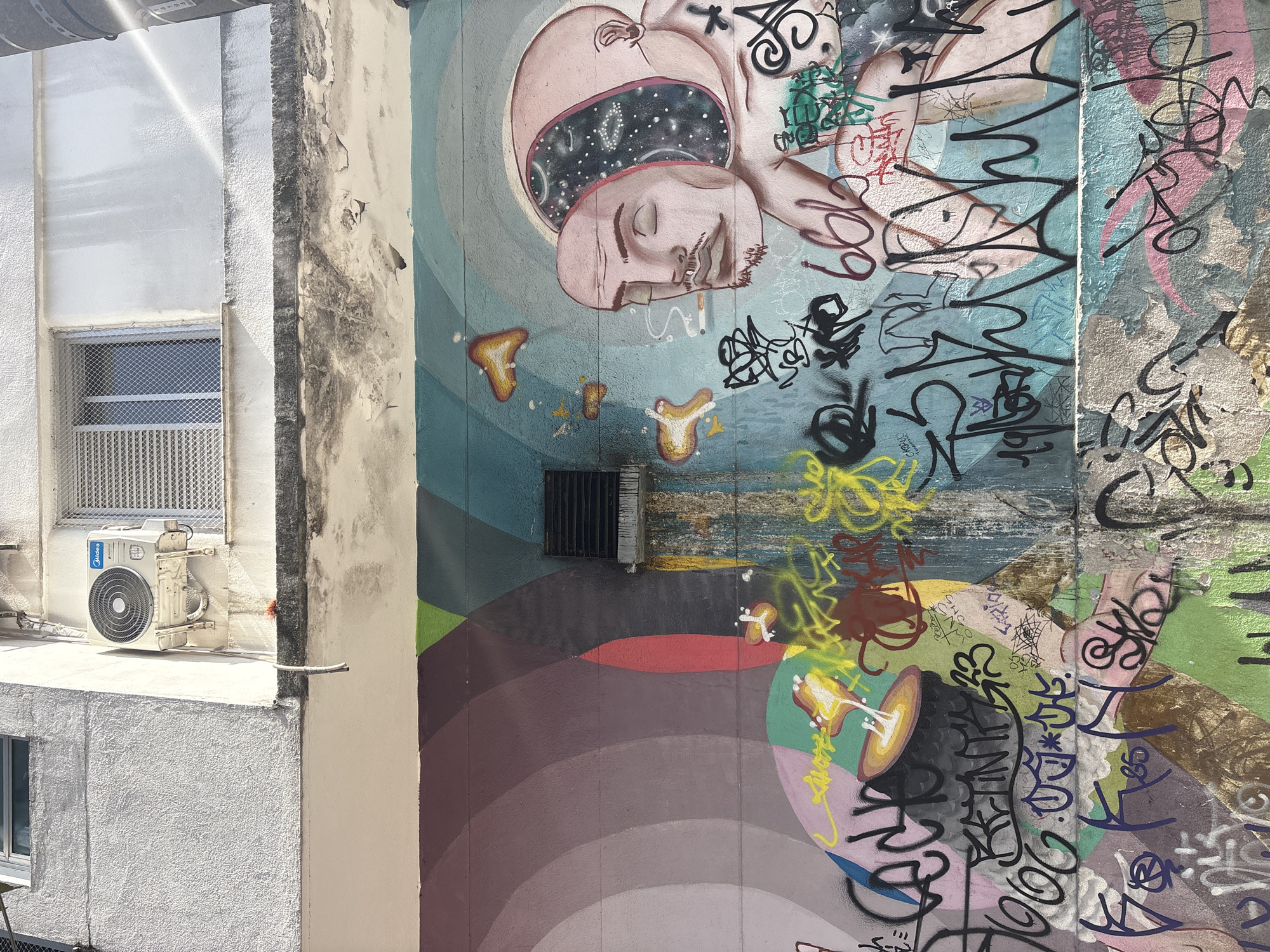 The image shows a mural on the exterior wall of a building in the city of São Paulo, Brazil. The mural depicts a surreal scene with a figure whose head is open, revealing a starry sky within. A detached human face, smoking a cigarette, is positioned in front of the figure's open head. Various abstract shapes and colorful patterns are also present on the wall. The lower portion of the wall is heavily covered in graffiti tags. An air conditioning unit is mounted on the wall above the mural, and a window with a metal grate is visible next to it. The building facade is textured and appears somewhat worn.