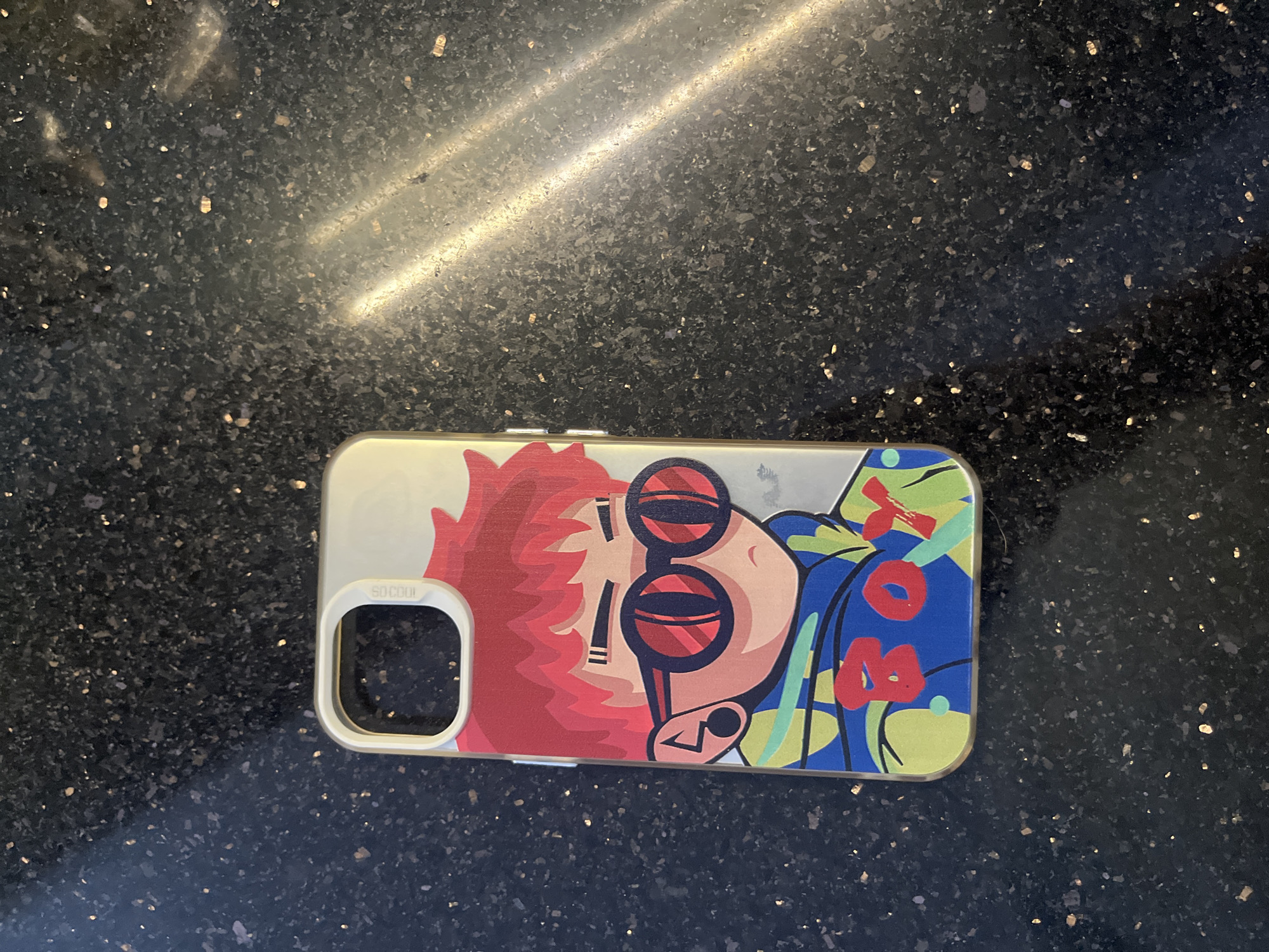 The image shows a cell phone case on a dark, speckled surface. The case has a cartoon graphic of a person with red hair and sunglasses. The background is dark and speckled, appearing to be a countertop. The location is not evident in the image.