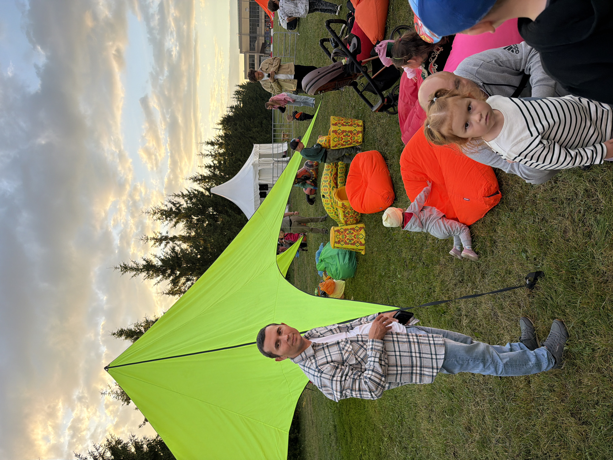 The image depicts an outdoor gathering, possibly a festival or park event, with a bright green tent dominating the left side of the frame. A man in a plaid shirt and jeans stands in front of the tent, holding a phone and a leash. The ground is covered in grass, with several large, colorful beanbag chairs and inflatable seating scattered around. In the background, a white tent is visible, along with trees and a building. Several people, including children and adults, are present, some interacting with each other and others appearing to relax on the beanbags. The sky is cloudy with hints of sunset colors.