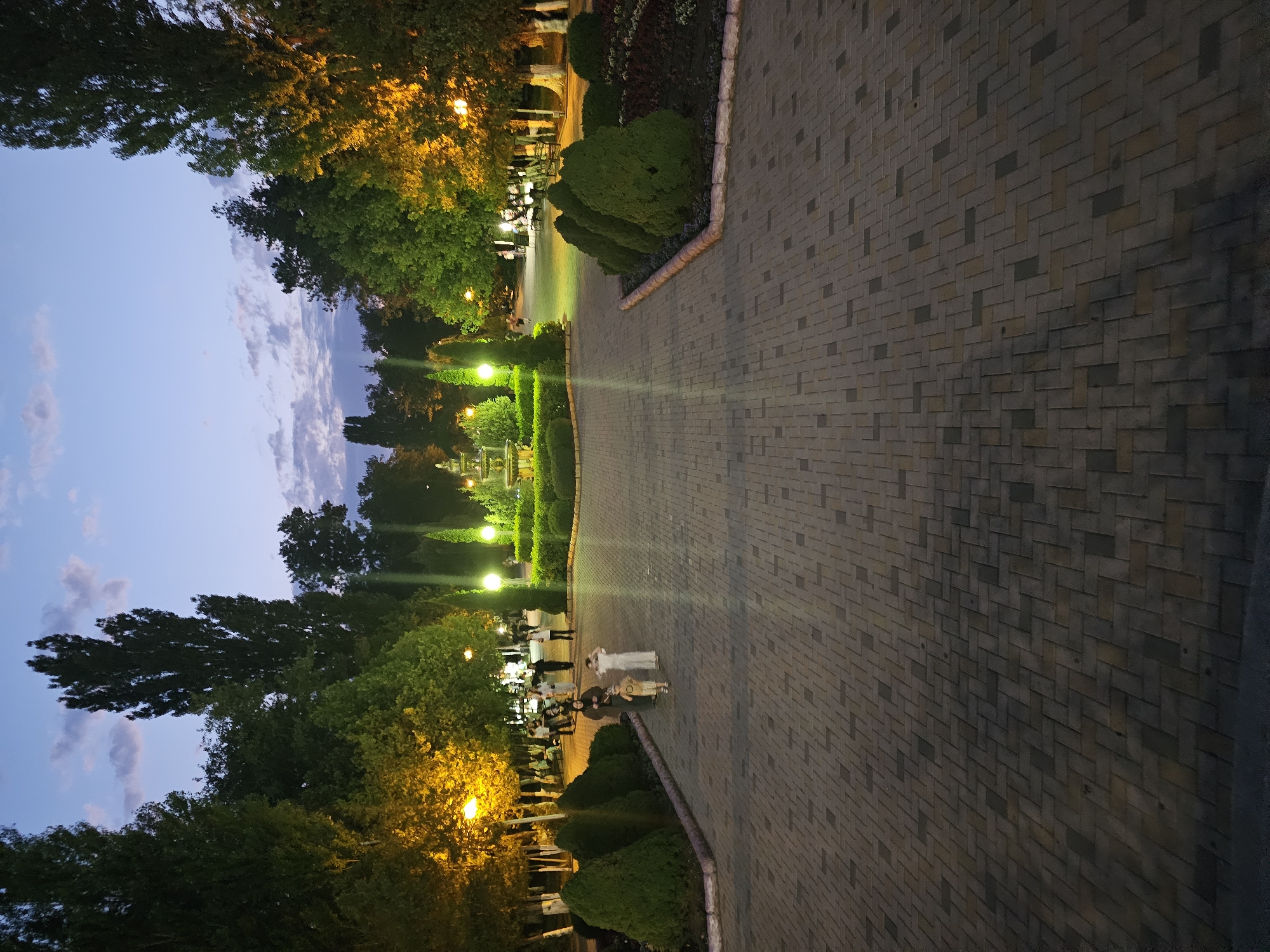 The image depicts a park pathway at dusk in Cherkessk, Russia. The pathway is paved with rectangular bricks laid in a herringbone pattern. On either side of the pathway, there are manicured bushes and flower beds. Trees line the edges of the park, some of which are illuminated by warm, yellow streetlights, creating a soft glow. In the distance, a fountain is visible, surrounded by more greenery. Several people are walking along the pathway, some appear to be families with children. The sky above is a deep blue, with scattered clouds, suggesting the transition from day to night. The overall atmosphere is serene and inviting.