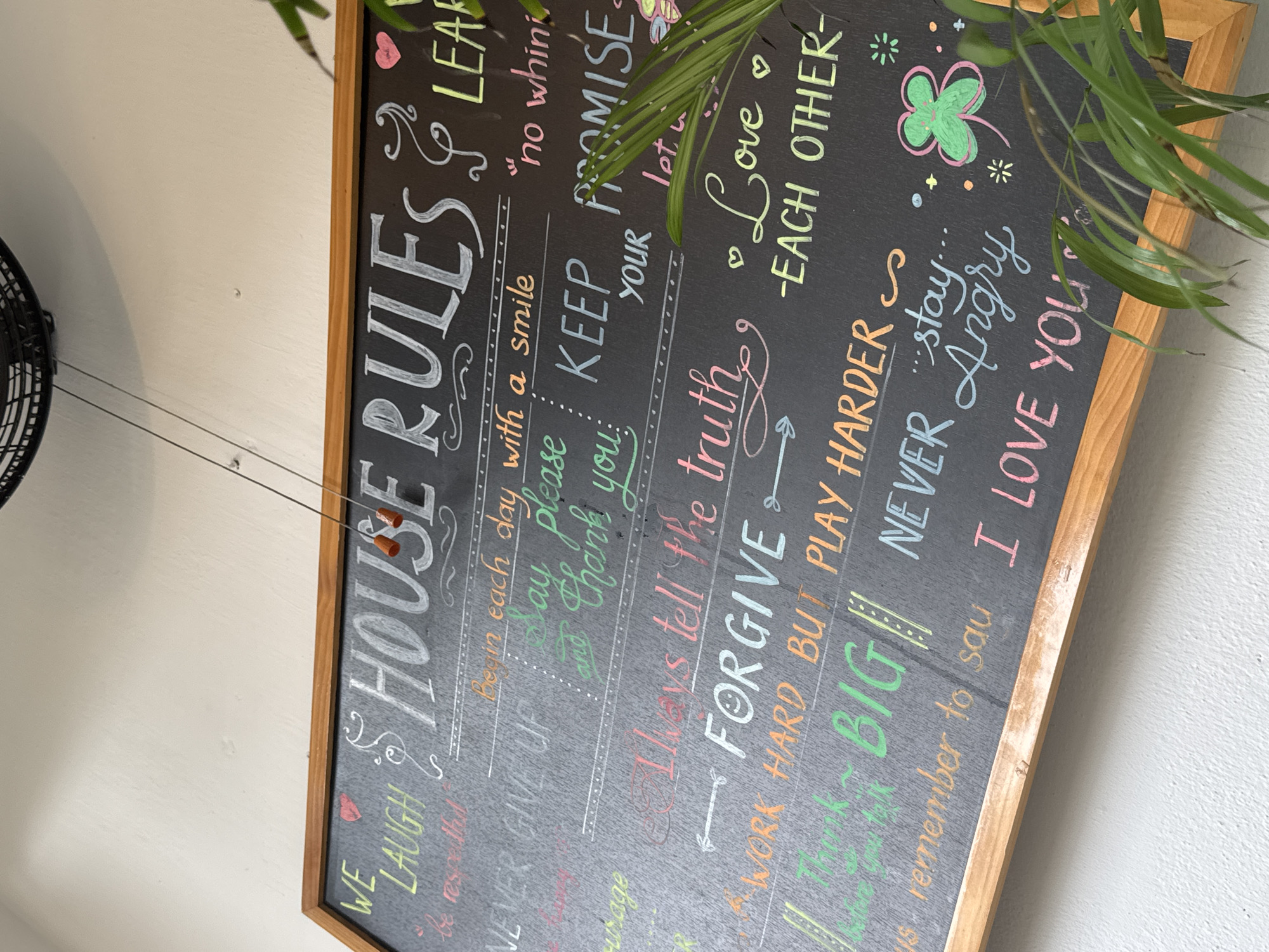 A framed chalkboard sign displaying "House Rules" in colorful chalk lettering hangs on a white wall. The sign features several rules, including "We Laugh," "Begin each day with a smile," "Keep your promise," "Love Each Other," and "I Love You." A green plant is partially obscuring the top right corner of the sign. A black fan is visible on the left side of the image. The scene appears to be indoors.