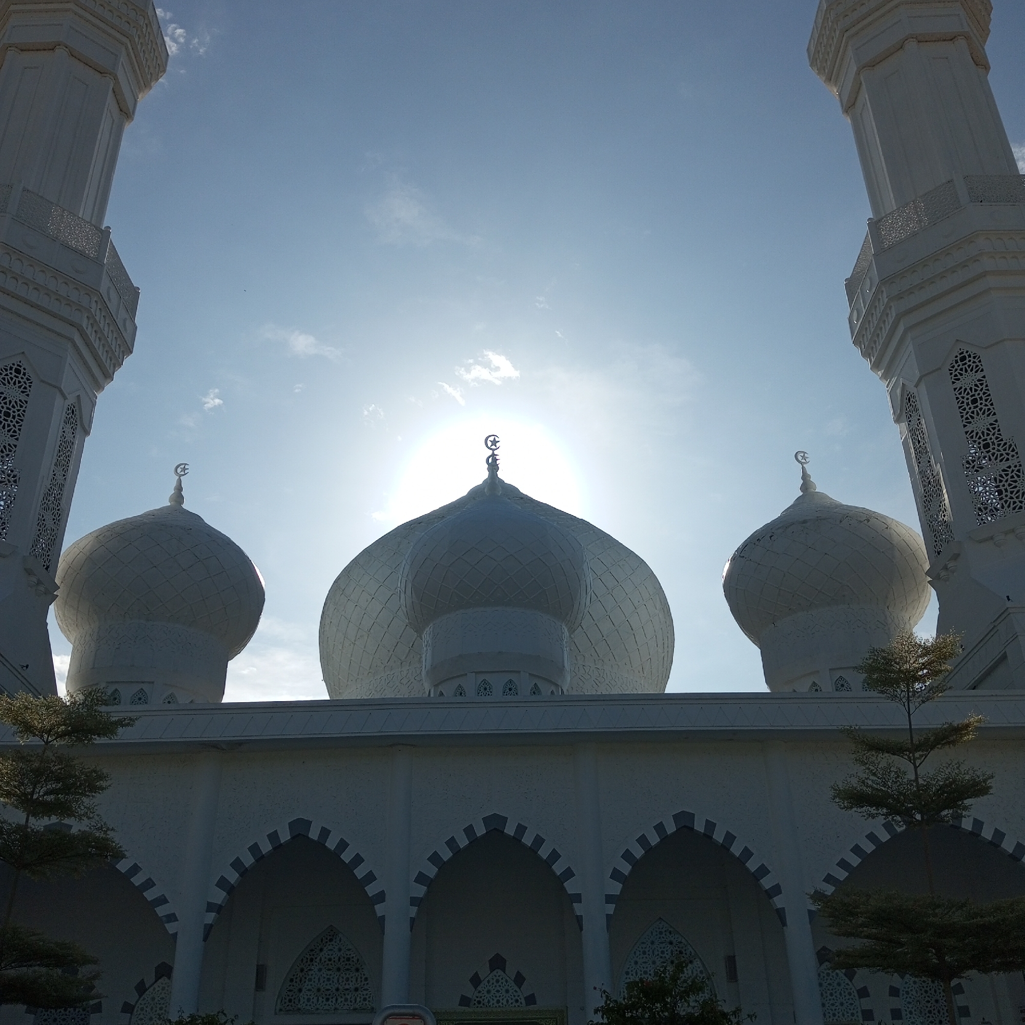The image captures a low-angle view of a mosque, likely in Meunasahaluedrienlhoksukon Barat, Indonesia, judging by architectural style. The mosque features white domes, with a larger central dome and two smaller ones flanking it. Each dome is topped with a crescent moon and star finial. Two tall, white minarets rise on either side, framing the domes. The architecture includes a series of arches and decorative patterns. The sun is positioned directly behind the central dome, creating a halo effect. Trees with green foliage are visible in the foreground, partially obscuring the base of the mosque. The sky is pale blue with scattered clouds.