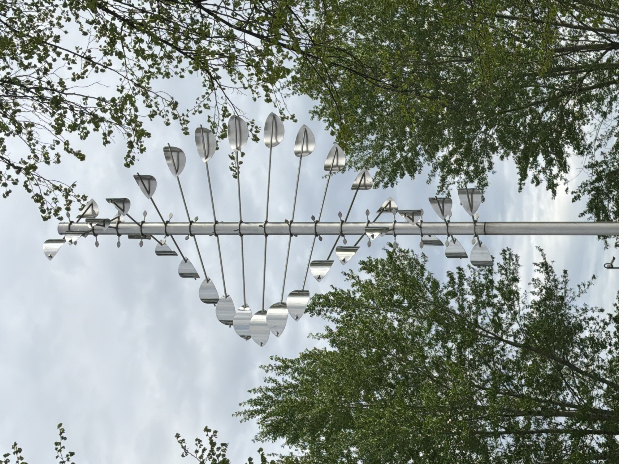 A low-angle shot depicts a horizontal metal pipe suspended between trees in Votkinsk, Russia. The sky is overcast. Affixed to the pipe are numerous metal rods, each holding a reflective, spade-shaped vane. The trees are green and leafy.