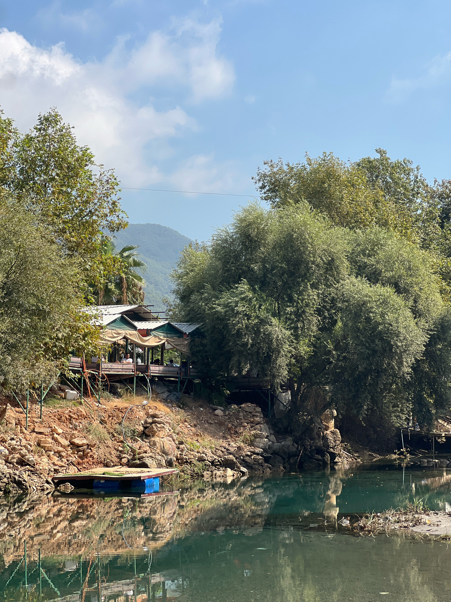 The image depicts a serene waterside scene with a restaurant built on stilts over the water. The restaurant has a white roof with green awnings and appears to be open-air, with tables and chairs visible. Lush green trees surround the structure, with a mountain visible in the background. A small blue and brown raft is floating on the water near the bank, which is rocky and covered with vegetation. The water is a clear, turquoise color and reflects the surrounding trees and the sky, which is partly cloudy. The overall impression is one of a peaceful, natural setting with a touch of rustic charm. This location is likely a riverside restaurant in Alanya, Turkey, possibly offering views of the Taurus Mountains.