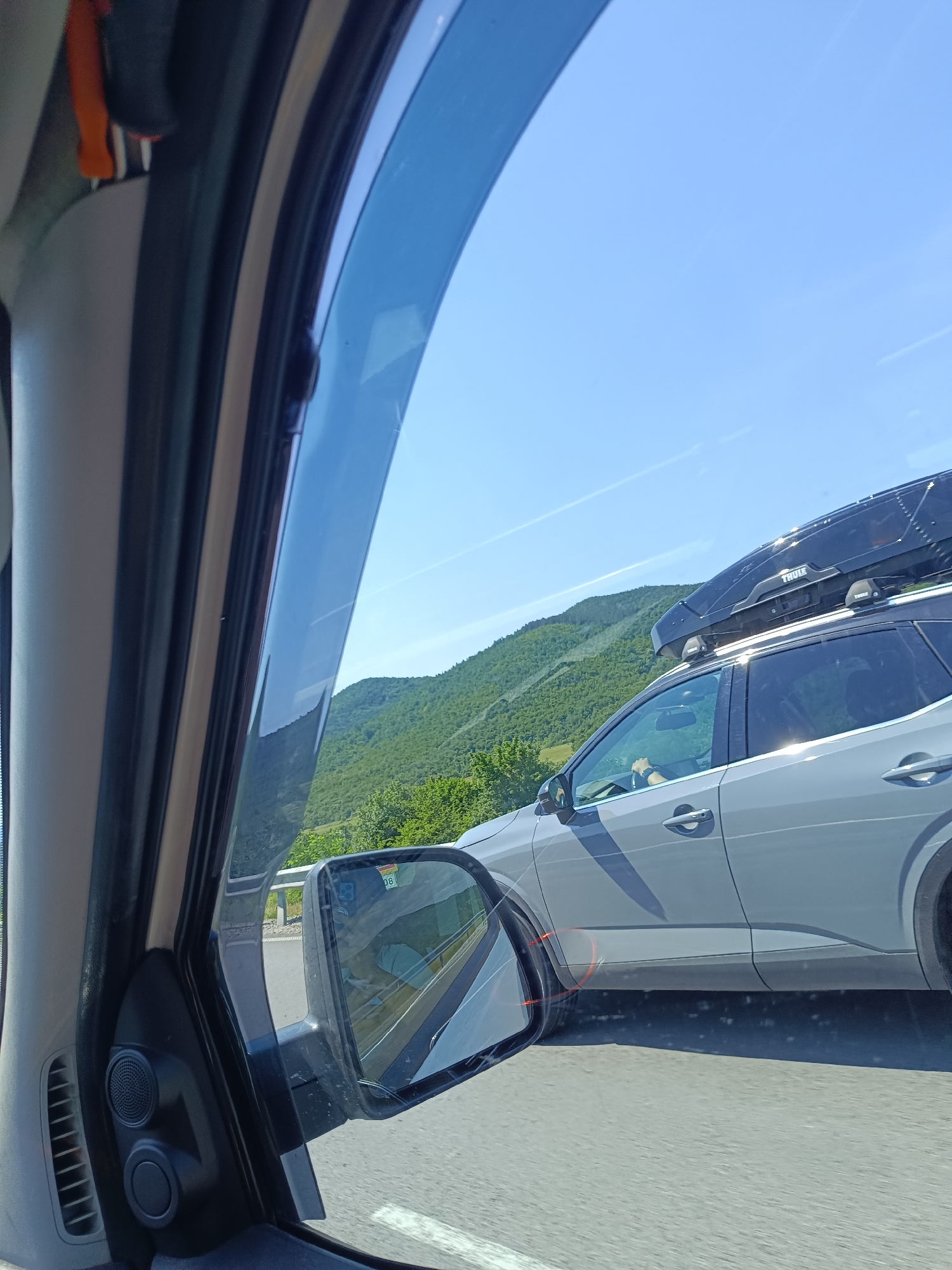 The image shows a view from the inside of a car, looking out the side window. The main subject is a gray SUV driving on a highway, visible in the side mirror and through the window. The SUV has a black cargo box on its roof with the brand name "THULE" visible. A person is driving the SUV, with their hand on the steering wheel. The background consists of lush green hills and a clear blue sky. The foreground shows the interior of the car, including part of the door panel, a speaker, and the car's side mirror. The highway is paved and appears to be in good condition. There are faint white lines on the road. The overall scene suggests a travel or road trip scenario.