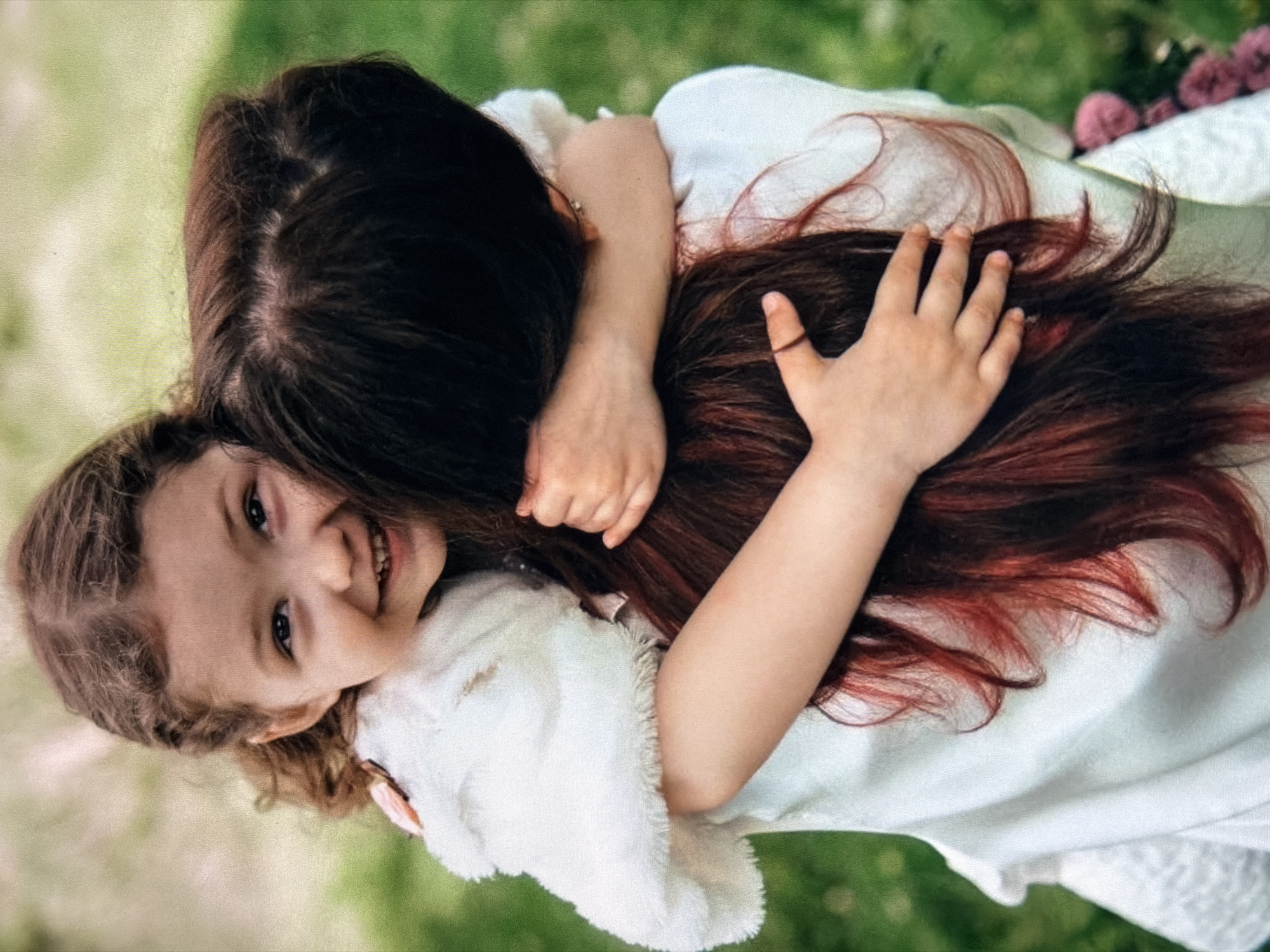 A close-up shot shows a young girl with curly brown hair embracing a woman from behind. The girl, with wide blue eyes and a joyful smile, is wearing a white fluffy jacket over a white dress. Her arms are wrapped around the woman's neck, and her hands rest on the woman's dark brown hair, which has reddish highlights. The woman's face is not visible, but her long, dark hair falls down her back. They are outdoors, with a blurred green background suggesting a grassy area. The lighting is soft, and the image has a shallow depth of field, emphasizing the subjects.