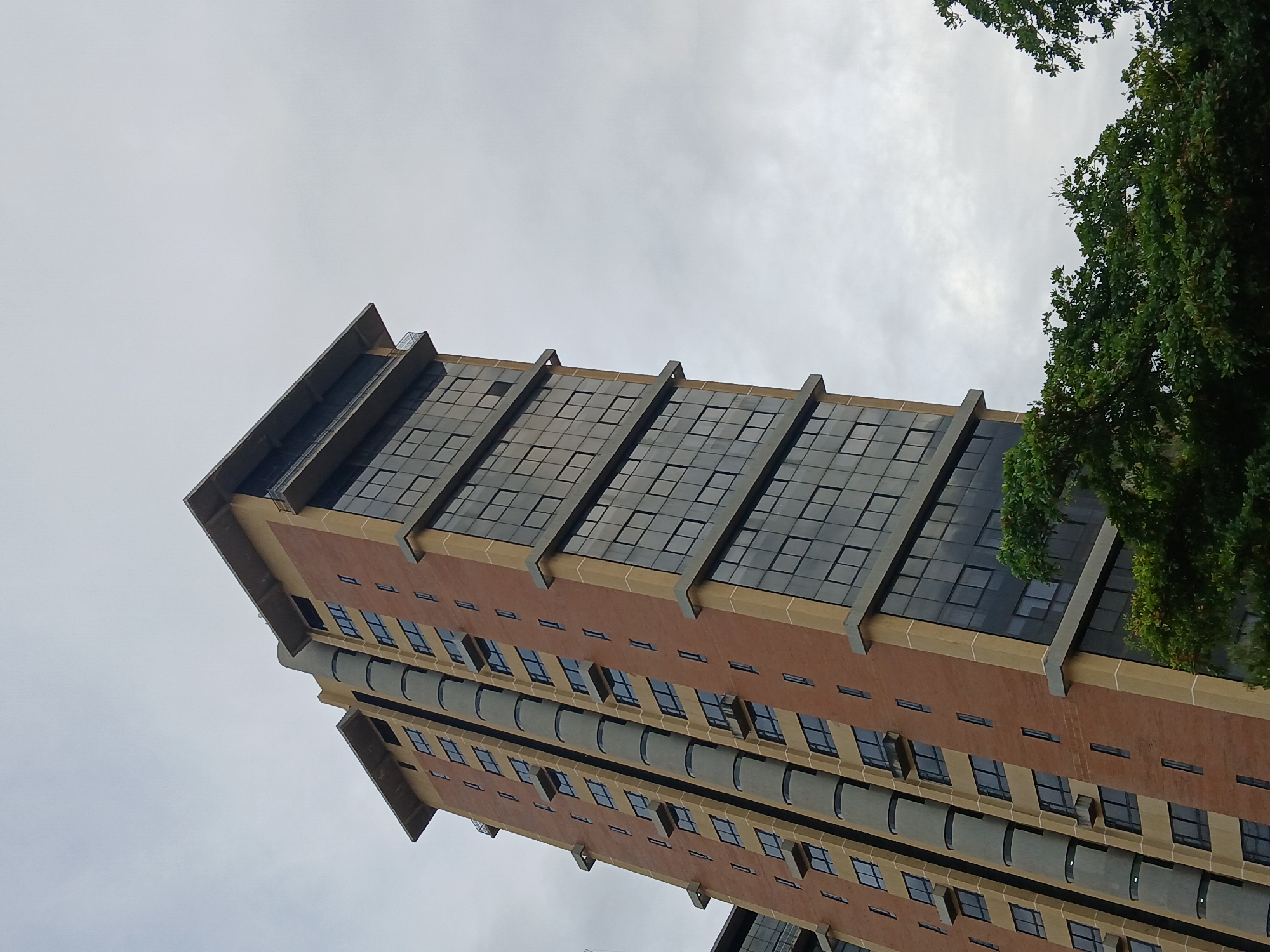The image displays the Torre Corporativa del Centro Comercial Ciudad Alta Vista II, a high-rise building located in Ciudad Guayana, Venezuela. The structure is observed from a low-angle perspective, emphasizing its verticality against an overcast, light gray sky that dominates the upper portion of the frame. The building's facade is characterized by alternating sections of dark, reflective glass windows and terra cotta-colored paneling, segmented by light beige horizontal and vertical bands. A distinctive gray, cylindrical column, composed of segmented elements, runs vertically along one side of the building. Smaller, dark rectangular windows and minor cantilevered projections are visible on the terra cotta sections. The building's apex features a flat roof with a substantial overhang and a recessed upper floor with a visible railing. Dense, dark green tree foliage is present in the lower right and bottom foreground, partially obscuring the building's base. No individuals or vehicles are depicted.