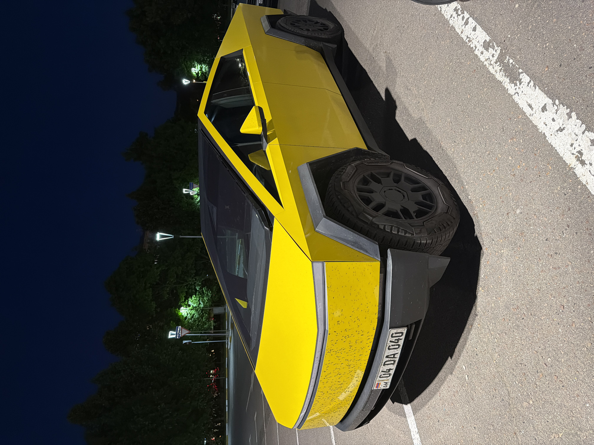 A vibrant yellow Tesla Cybertruck is captured at night in an outdoor parking lot. The vehicle is positioned diagonally, with its rear-right side prominently featured in the foreground. Its distinctive angular body is painted a bright yellow, contrasting sharply with the black trim and dark, rugged wheels. A spare tire is visibly mounted on the rear.
The surface of the Cybertruck has numerous small droplets or dust specks, particularly noticeable on the rear panels. The license plate, "AM 04 DA 040," is clearly visible on the rear, indicating an Armenian registration, though the contextual location is Moscow, Russia.
The setting is an asphalt surface, with a white parking line partially visible on the right. The time of day is nighttime, characterized by a dark blue sky and the illumination provided by several modern streetlights in the background. Dark trees line the background, partially lit by the streetlights, which cast a bright glow. The scene is quiet, with no people or other vehicles immediately visible.