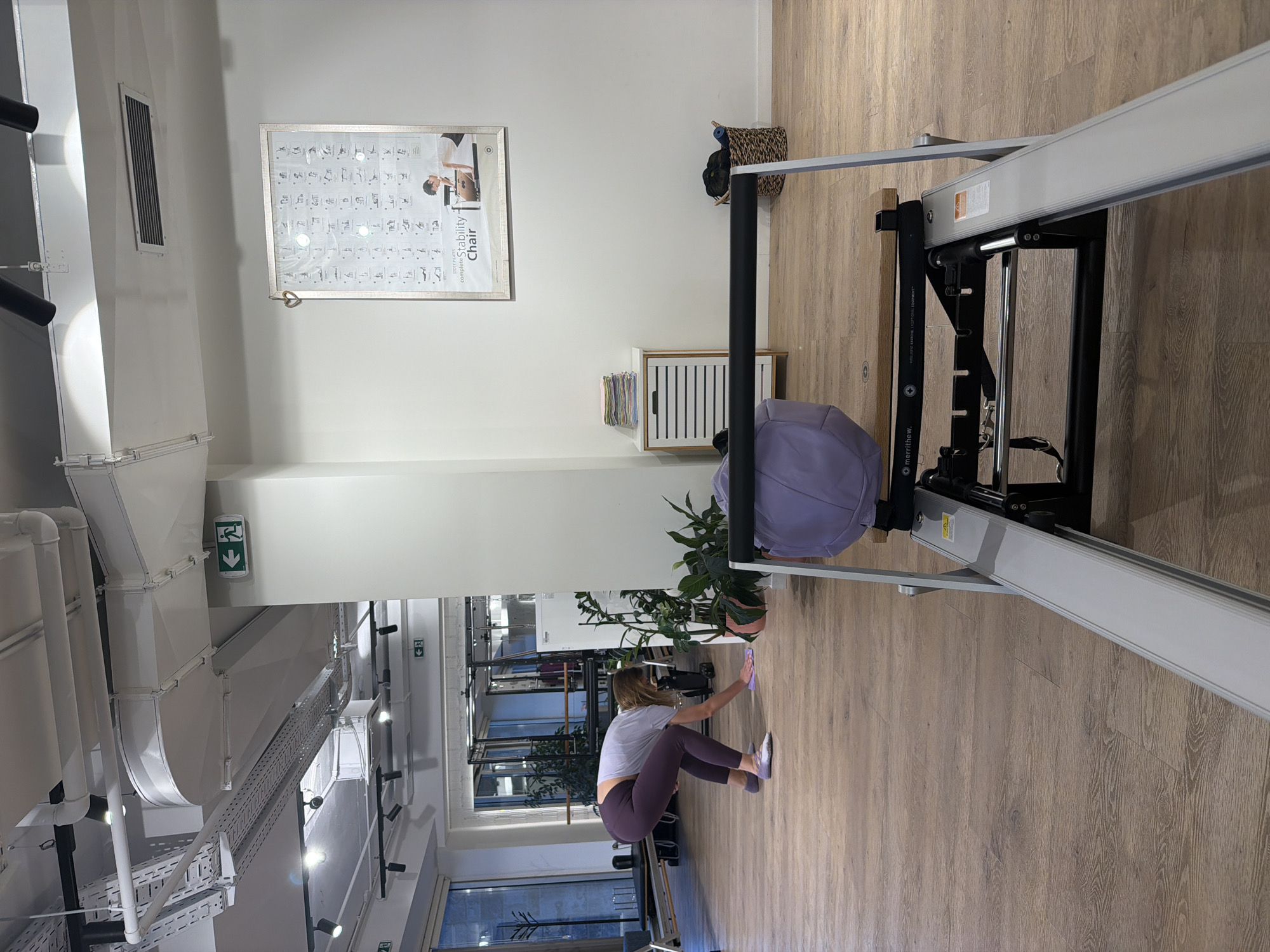 The image depicts the interior of a Pilates studio. In the foreground, a Pilates reformer machine is visible, angled towards the viewer. To the left, a woman wearing purple leggings and a grey top is on her hands and knees on the wooden floor, wiping it with a purple cloth. In the background, other Pilates equipment, including reformers, is set up. A tall plant stands near the woman, and a small white cabinet with drawers is to the right of the plant. On the wall above the cabinet, a framed poster displays various Pilates exercises. The room has a high ceiling with exposed industrial-style ventilation ducts and lighting fixtures. The overall environment suggests a clean, modern fitness space. The location in Boreti, Montenegro is not indicated by any visual cues in the image.