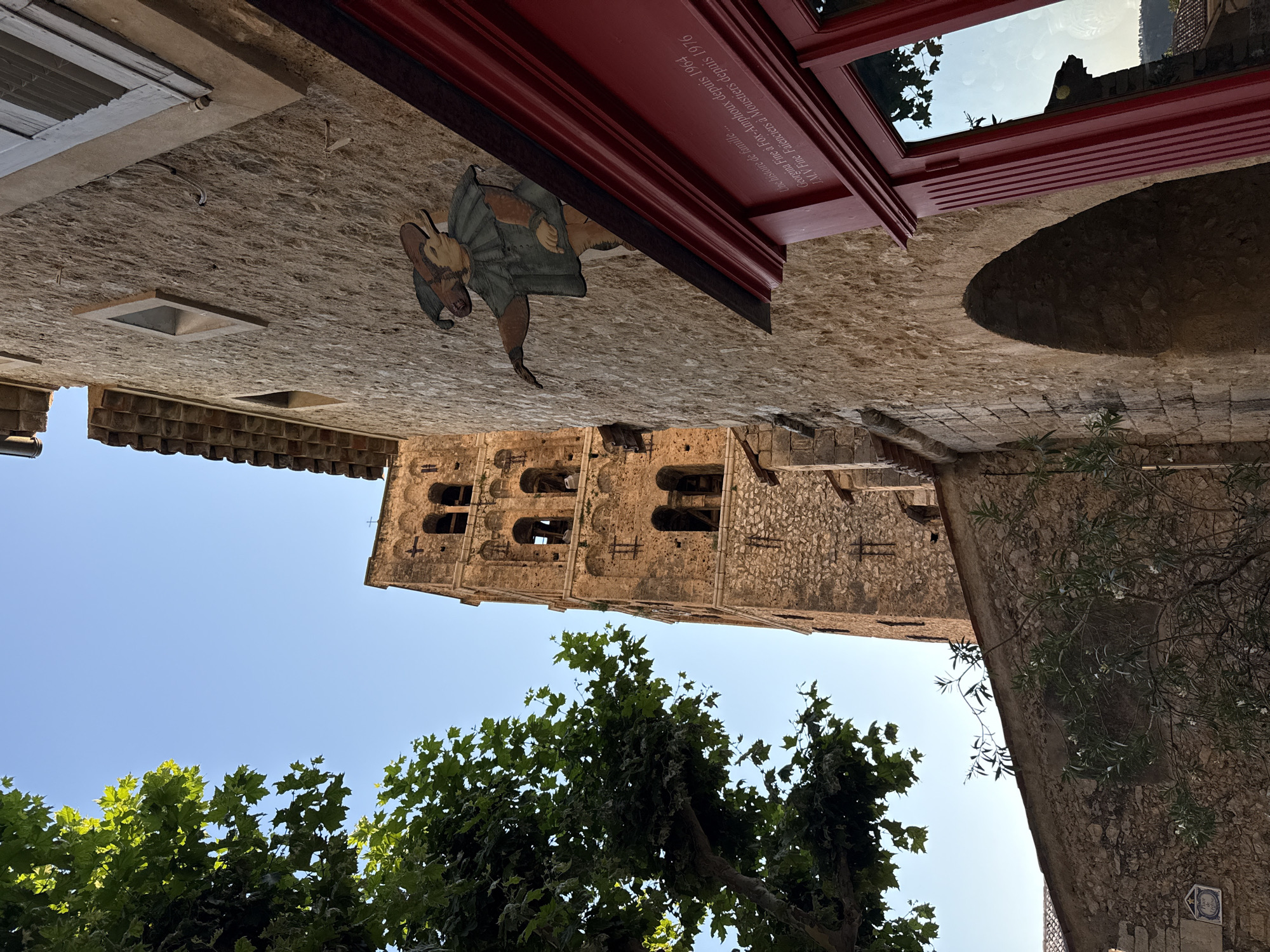 The image depicts a narrow street scene in Moustiers-Sainte-Marie, France, with a tall, ancient stone bell tower dominating the background. In the foreground, a weathered stone building occupies the right side, featuring an arched entryway and a prominent red-trimmed window. A painted wooden sign of a jester figure is affixed to the building's facade. Inscriptions are visible on the red trim of the window. A large tree with green leaves is situated to the left, partially obscuring the view of the bell tower and a portion of the sky. The overall scene suggests a historic, possibly medieval, European town.
