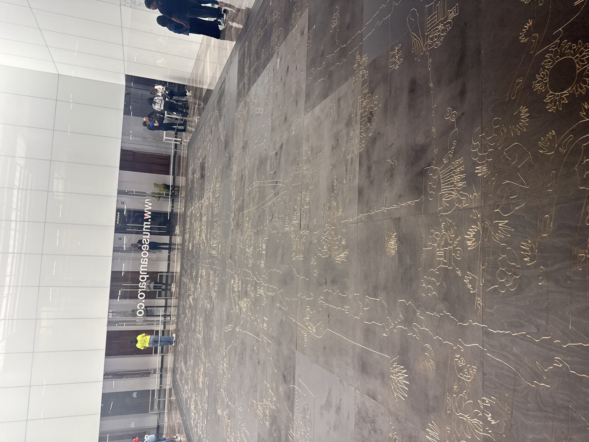 The image depicts a wide, tiled floor area with etched designs resembling indigenous Mexican art, possibly glyphs and depictions of people, plants, and celestial objects. The tiling extends from the base of a large glass window wall that reflects people inside the building. A website address “www.museosobsto.com” is visible on the glass. Several people can be seen inside and outside the building, reflected or standing in the vicinity. The perspective of the image suggests it was taken at or near the floor level, focusing on the artwork. Given the website address, the location is likely the Museo de la Constancia Mexicana in the city of Puebla, Mexico.
