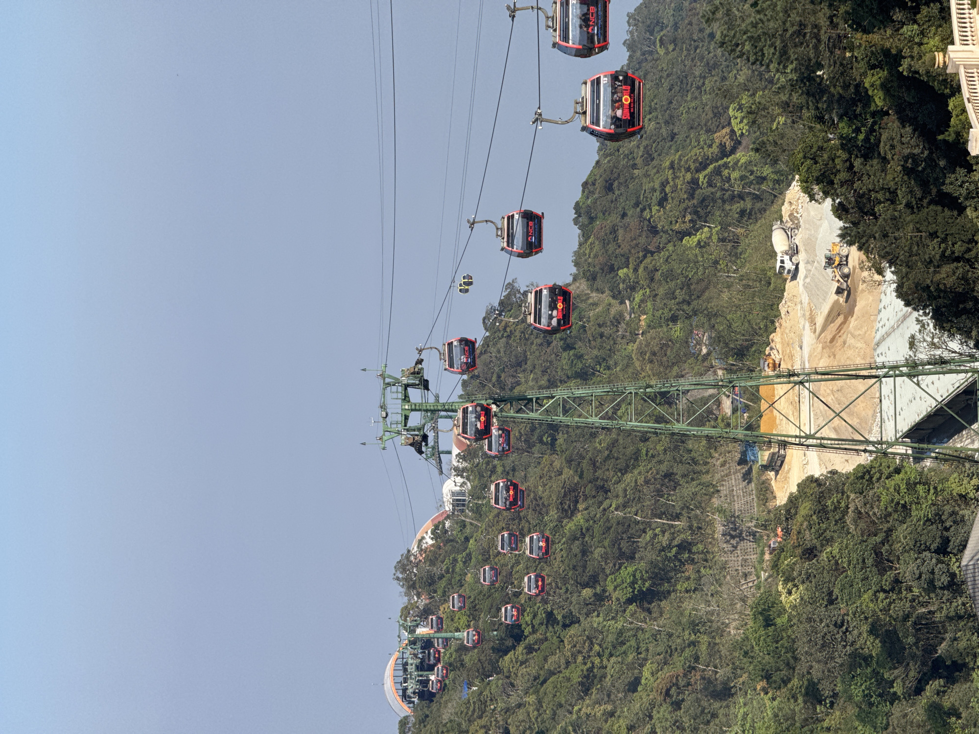 The image depicts a panoramic view of the SunWorld Ba Na Hills cable car system traversing a densely forested mountainous area west of Đà Nẵng, Vietnam. The upper portion of the frame features a clear, pale blue sky.
Numerous red and black cable car gondolas, approximately 20-30 in count, are suspended from multiple overhead steel cables. Many gondolas display "SUNWORLD BA NA HILLS" branding in white and red text, along with a "NCB" logo. Indistinct human figures are visible inside several gondolas, indicating passenger transport. The gondolas are distributed across the frame, suggesting active operation.
The cable system is supported by several green-painted metal lattice towers. A prominent tower occupies the foreground, centered vertically, extending from the bottom of the frame upward. In the background, on distant forested hilltops, various modern architectural structures with arched roofs and domes are visible, consistent with a resort complex.
In the lower-midground, a cleared construction site disrupts the natural forest cover. This site features excavated earth and piles of aggregate. Identifiable heavy machinery includes a white concrete mixer truck, a yellow front-end loader, an orange excavator positioned on a terraced slope, and a dark blue dump truck. A terraced concrete retaining wall with visible grid reinforcement is present on a slope within the construction zone, alongside a large, flat concrete slab or foundation. The surrounding terrain is characterized by lush, verdant evergreen forest.