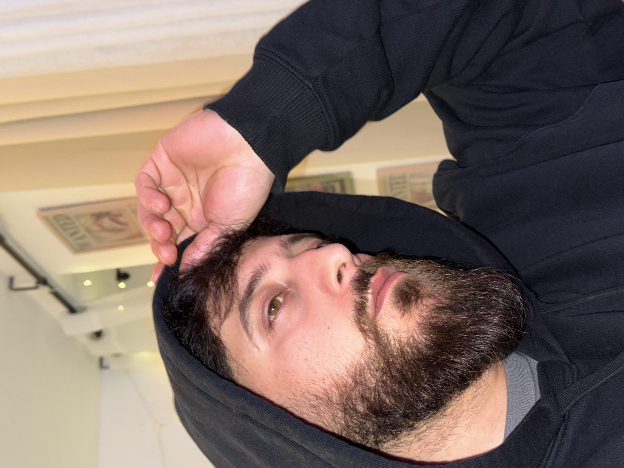 The image presents a close-up, upper-body shot of a male subject within an indoor setting in Strasbourg, France. The man has a full dark brown beard and mustache, dark hair partially visible beneath a black hood, and light brown eyes. He is wearing a black hooded sweatshirt over a grey crew-neck shirt. His right hand is raised, with fingers resting on his forehead, partially adjusting the hood. His gaze is directed towards the upper right quadrant of the frame. The background consists of a light-colored wall. A framed 'WANTED' poster is visible on the wall to the upper left, and a strand of decorative string lights hangs across the top. Light-colored curtains are visible on the right side of the frame.