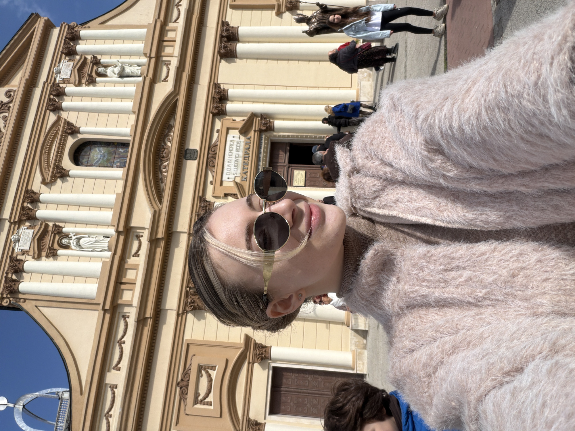 A woman wearing round sunglasses and a fuzzy pink coat stands in front of the Cathedral of St. Louis in Plovdiv, Bulgaria. The cream-colored facade of the cathedral features classical architectural elements such as columns, arches, statues, and ornate carvings. People are seen walking in front of the cathedral, and the sky is blue.