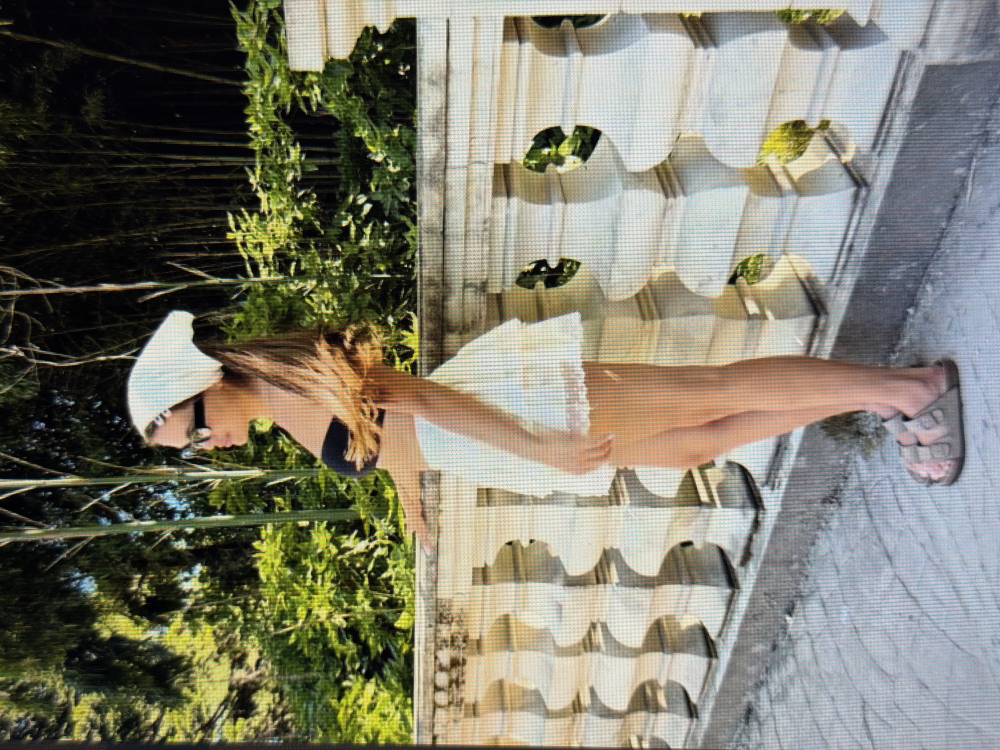 A person is standing on a paved walkway next to a white decorative railing. The person is wearing a white cap, sunglasses, a black bikini top, and a white skirt. They have long brown hair and are wearing sandals. The person is facing left and appears to be looking towards the left. Behind the person and the railing is a lush green area with trees and bamboo. The scene appears to be outdoors during the daytime, likely on a sunny day given the shadows and bright lighting. The overall mood seems relaxed and summery. There is no visible text in the image. The location context is Sirius, Russia.