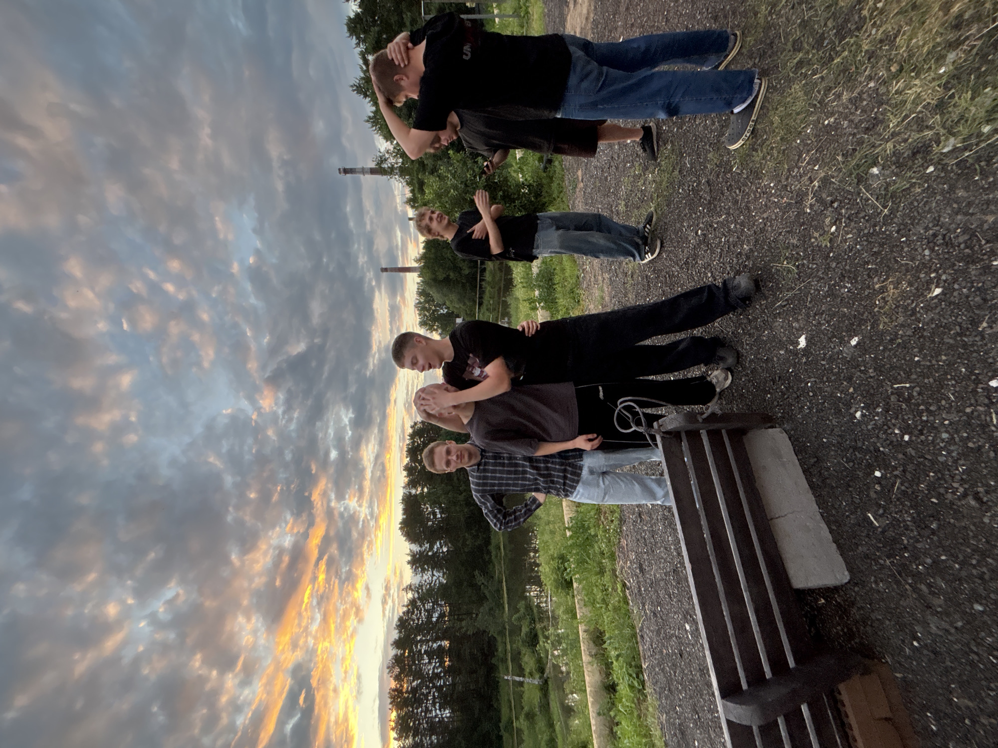 In Chaikovsky, Russia, a group of young men stand outdoors near a body of water and a line of trees. The sky is filled with dramatic clouds, tinged with orange and yellow from the setting sun. Two of the men are positioned in the foreground, one with his arm around the other and their hands obscuring his face. To their left, a man in a plaid shirt and jeans stands with his hands on his hips. Further back, three more young men are gathered, one looking at a phone. A dark wooden bench sits in the lower left corner of the frame. The ground is covered in dark gravel and sparse grass. In the distance, two industrial chimneys are visible against the twilight sky.