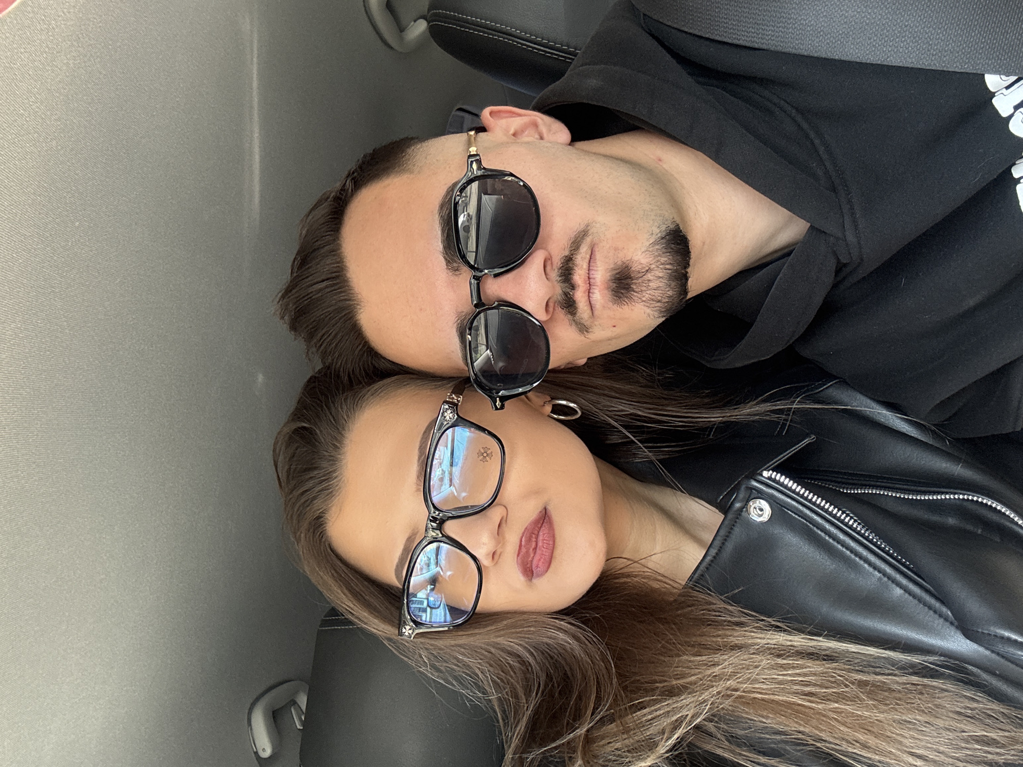 A selfie-style photograph shows a man and a woman posing together in what appears to be the interior of a car. The image is oriented horizontally, with the individuals' heads tilted towards each other.
The man is positioned at the top of the frame, wearing a black hoodie and dark sunglasses. He has short, dark hair and a small goatee. The woman is located below him, with long, light brown hair and wearing glasses with transparent lenses. She is dressed in a black leather jacket.
The background shows the interior of the car, including the headrest of a seat and the ceiling lining, which appears to be gray.
There is no specific indication of the location being Sofia, Bulgaria. Therefore, I cannot confirm the location in the city of Sofia, Bulgaria.
