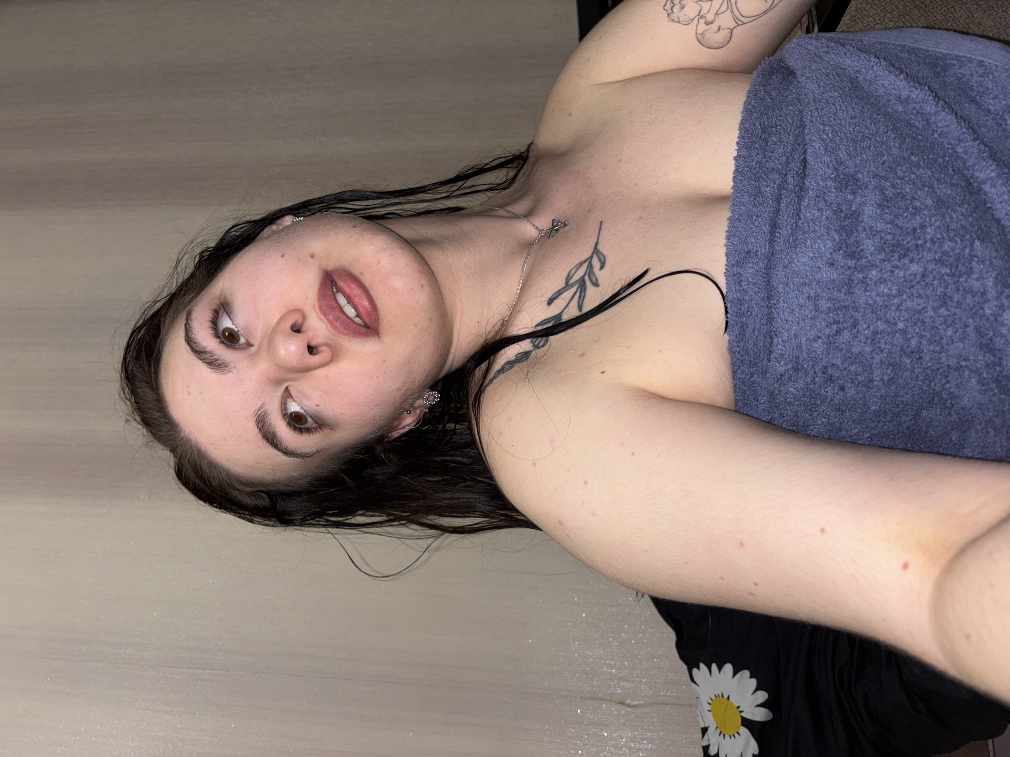 A medium shot depicts a woman with damp, dark hair, light skin, and brown eyes. She is partially covered by a blue towel, with a visible portion of a black shirt with a white daisy print at the bottom. A silver necklace adorns her neck. Tattoos are visible on her upper chest and arm. The background consists of a light brown surface, resembling a floor or wall. The image appears to be taken indoors under artificial lighting. The location, Perm, Russia, is not directly indicated in the image.