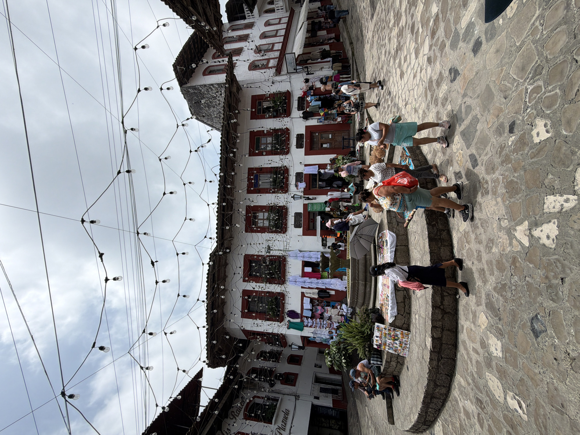 The image depicts a bustling scene in the city of Cuetzalan, Mexico, possibly in a central plaza. A multi-story building with white walls and red-trimmed windows dominates the background. The facade is adorned with hanging plants, and clotheslines are strung between windows, displaying various items. Numerous people are visible, some browsing items displayed on tables or blankets set up as makeshift market stalls. Others are walking through the plaza. The ground is paved with irregular stone blocks. Above, a network of wires with hanging light bulbs crisscrosses the space. The sky is overcast, suggesting diffused daylight.