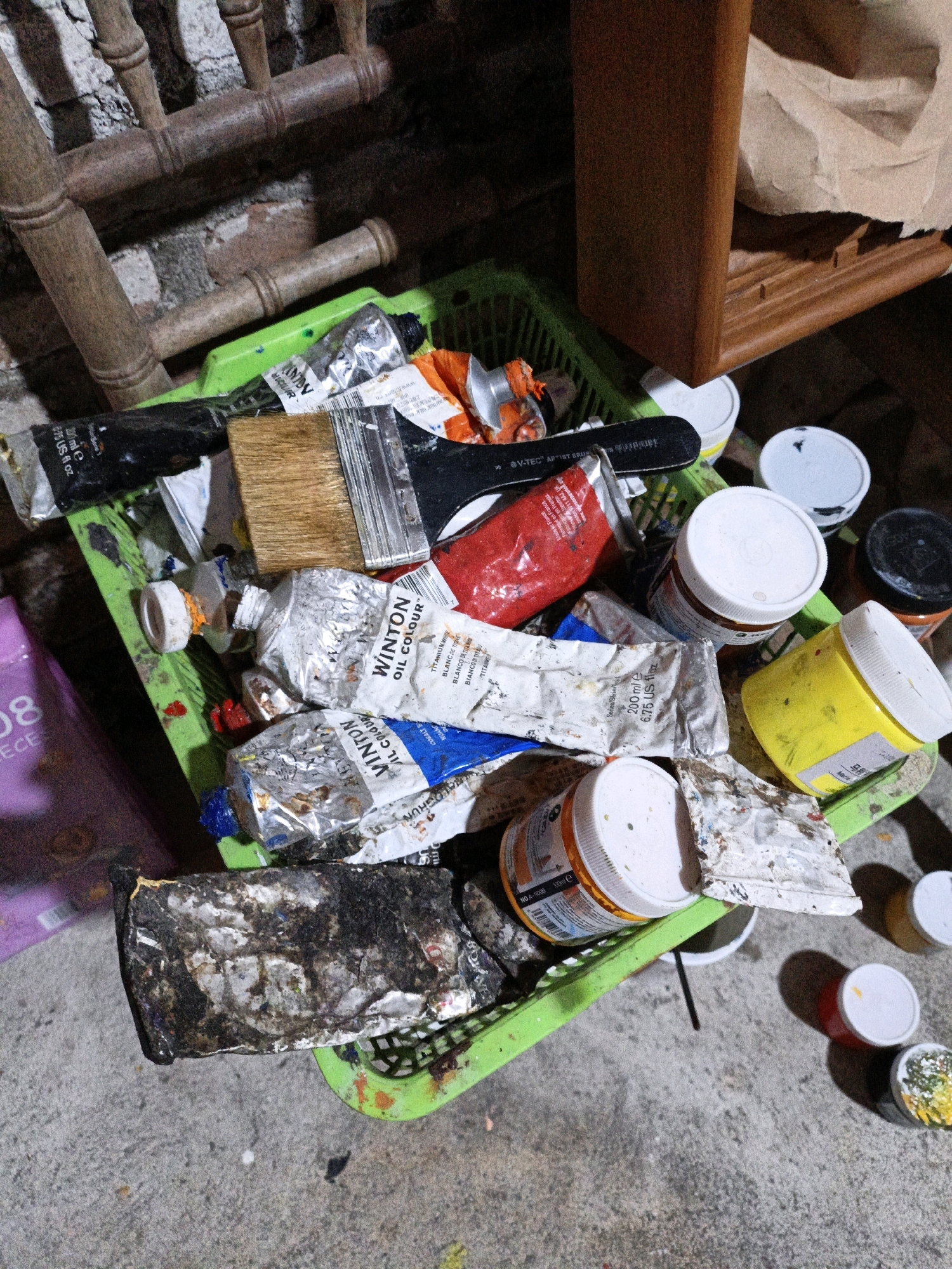 The image depicts a close-up, overhead view of a green plastic basket filled with art supplies, primarily tubes of paint and brushes. The basket sits on a concrete floor, likely in a storage area or workspace. The paint tubes are in various states of use, with some appearing crushed and others relatively intact. Several brushes are visible, some with paint residue on their bristles. Various containers of paint, including jars with white lids, are also present in and around the basket. In the background, parts of wooden furniture and a brick wall can be seen. The lighting is dim, creating shadows. There is no indication of the city of Cikal, Indonesia, or any specific location within it. The scene appears to be a still life of art materials, possibly in a state of disarray.